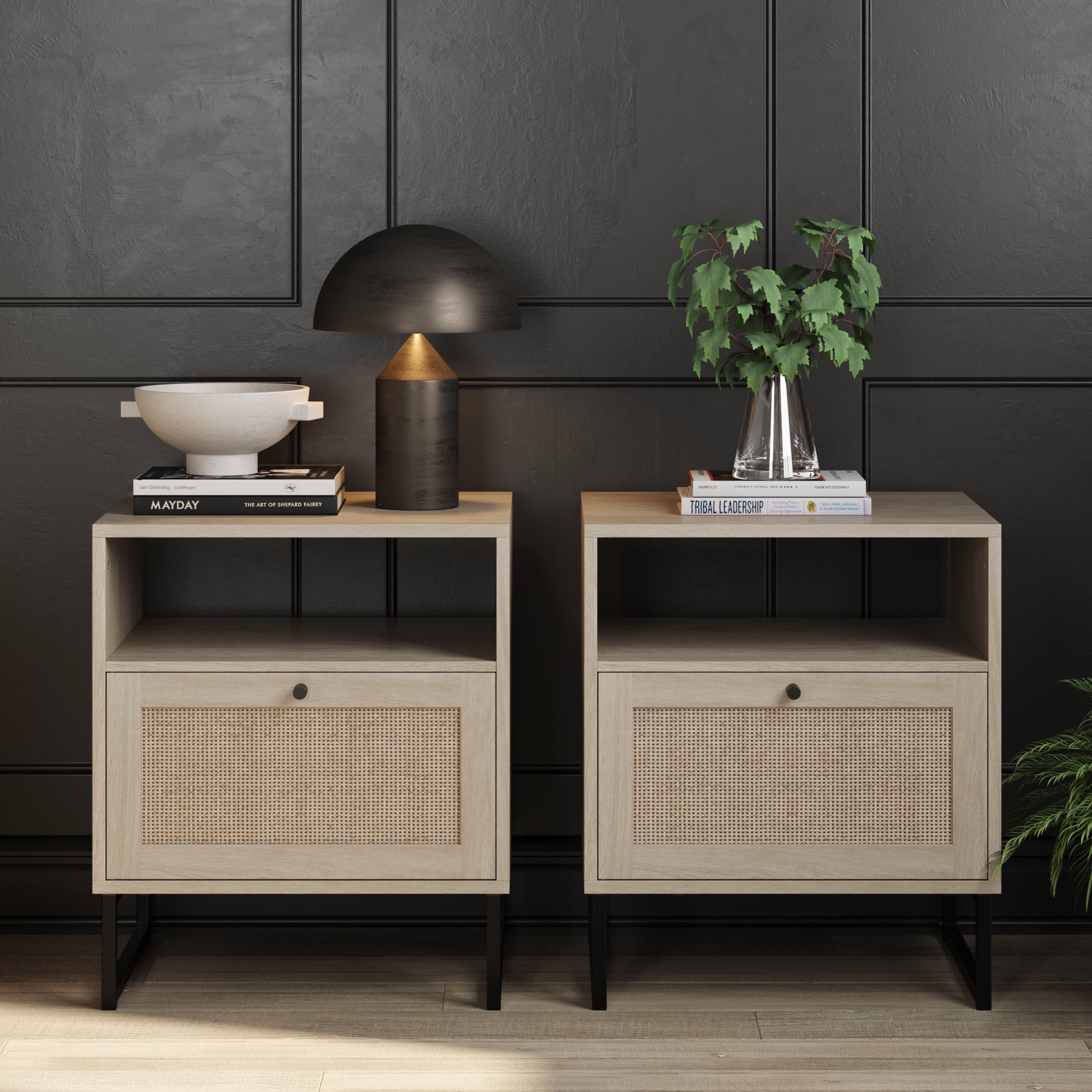 Two modern wooden nightstands with cane-front drawers sit against a dark paneled wall. One holds books, a bowl, and a black lamp; the other has books and a potted plant. Both have black metal legs and a light wood finish.