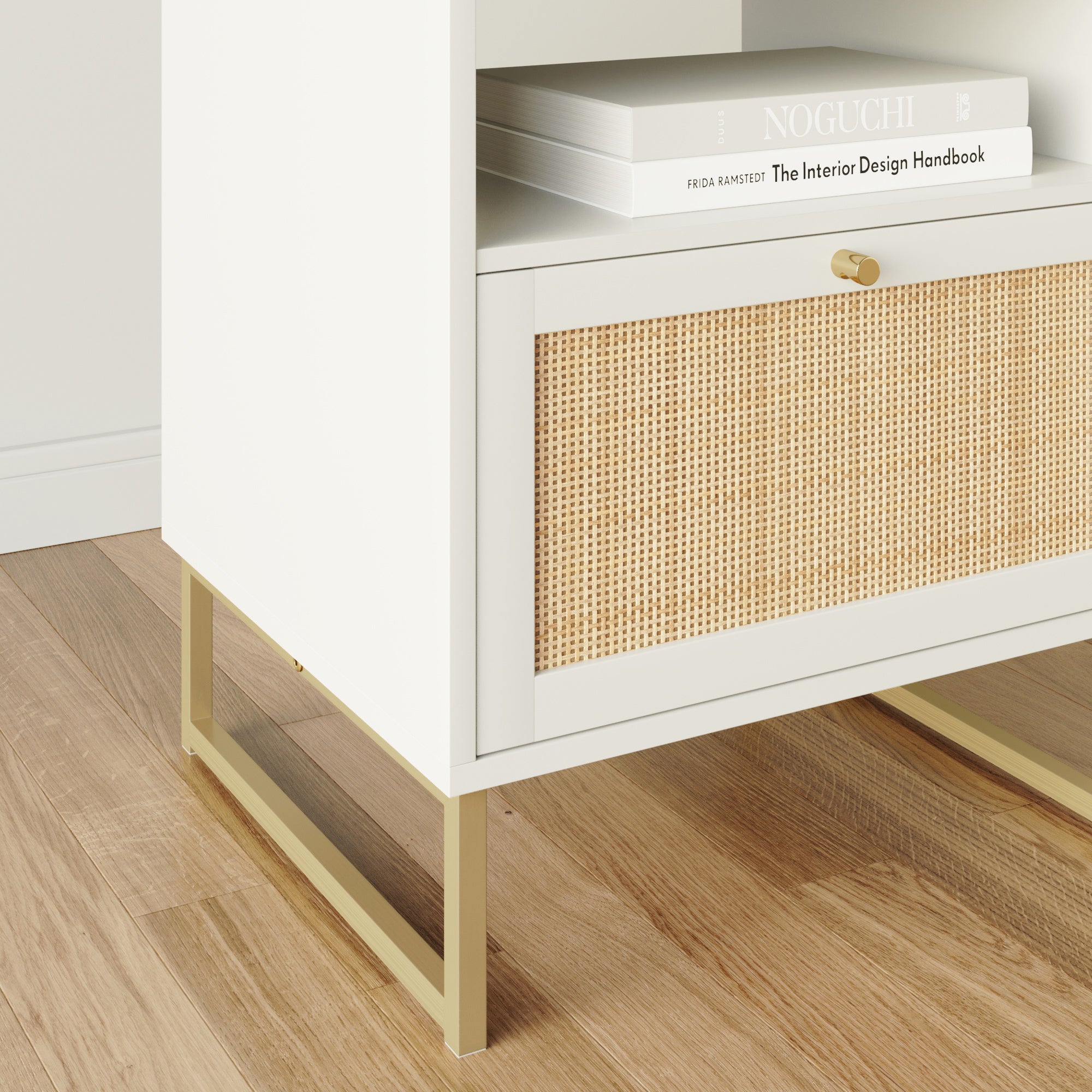 A white modern cabinet with gold legs stands on a wooden floor. The cabinet features a woven rattan drawer and has a book titled The Interior Design Handbook placed on top.