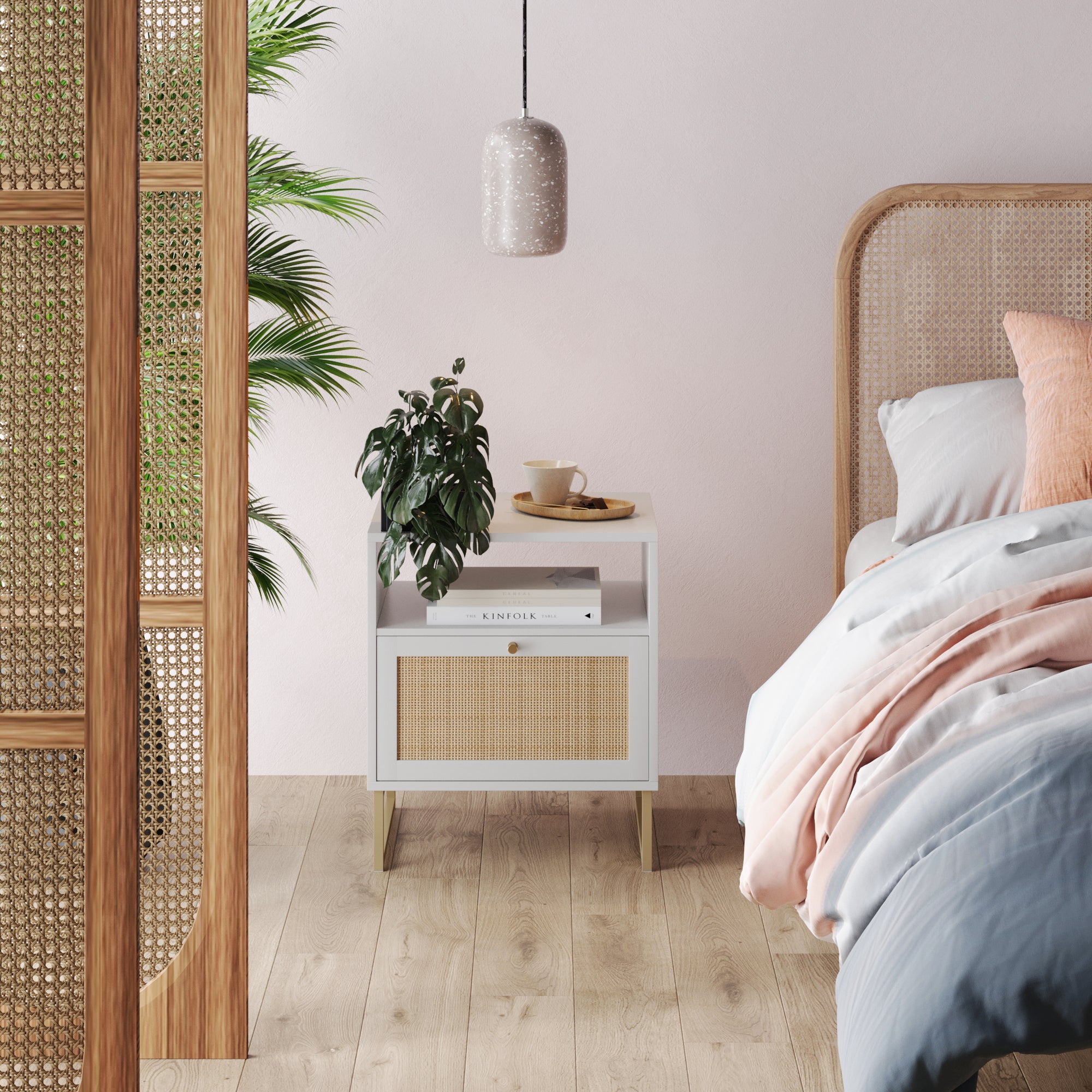 A cozy bedroom corner with a rattan bed, white nightstand, potted plant, book, cup, and pendant light against a pale pink wall. Wooden floors and a partition with rattan detail complete the calm setting.