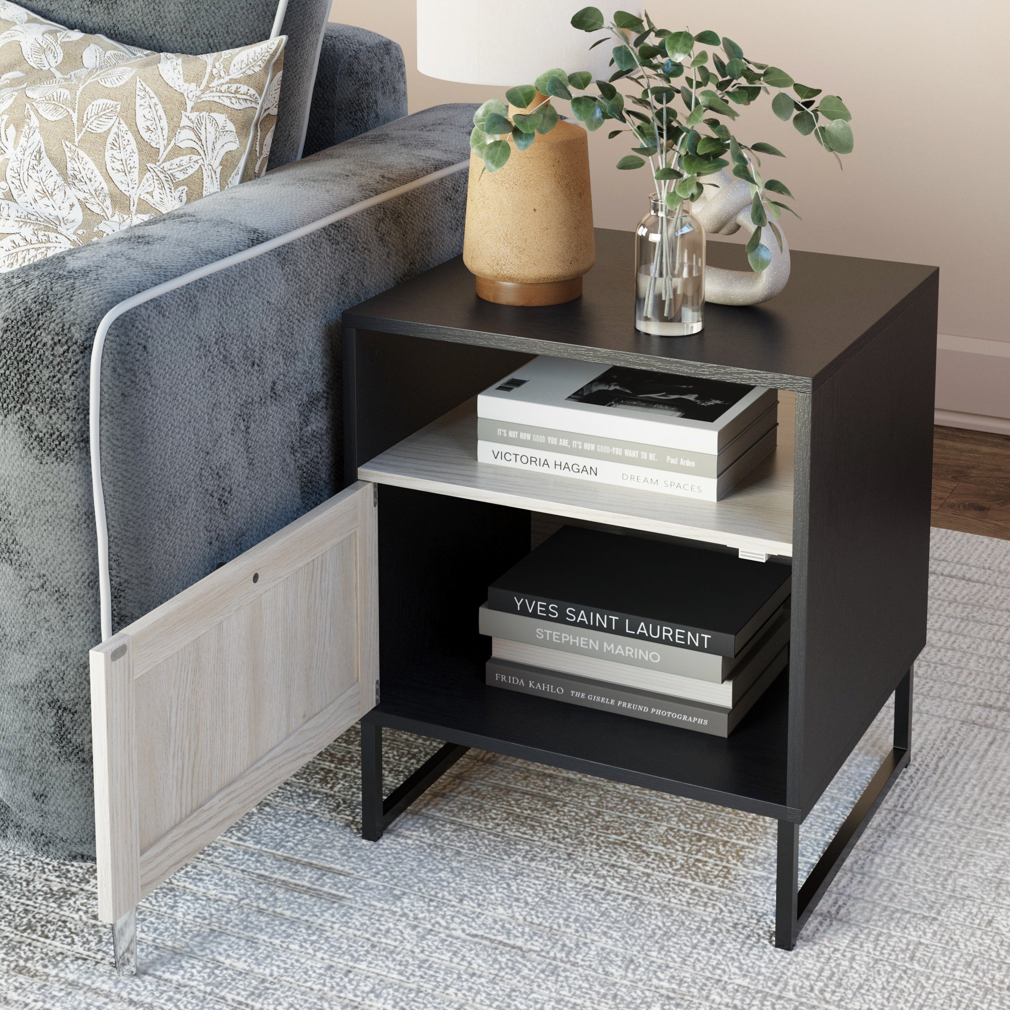 A modern black side table next to a gray sofa holds stacked books, a beige lamp, and a glass vase with greenery. One lower cabinet door is open, revealing more books on the shelf inside.