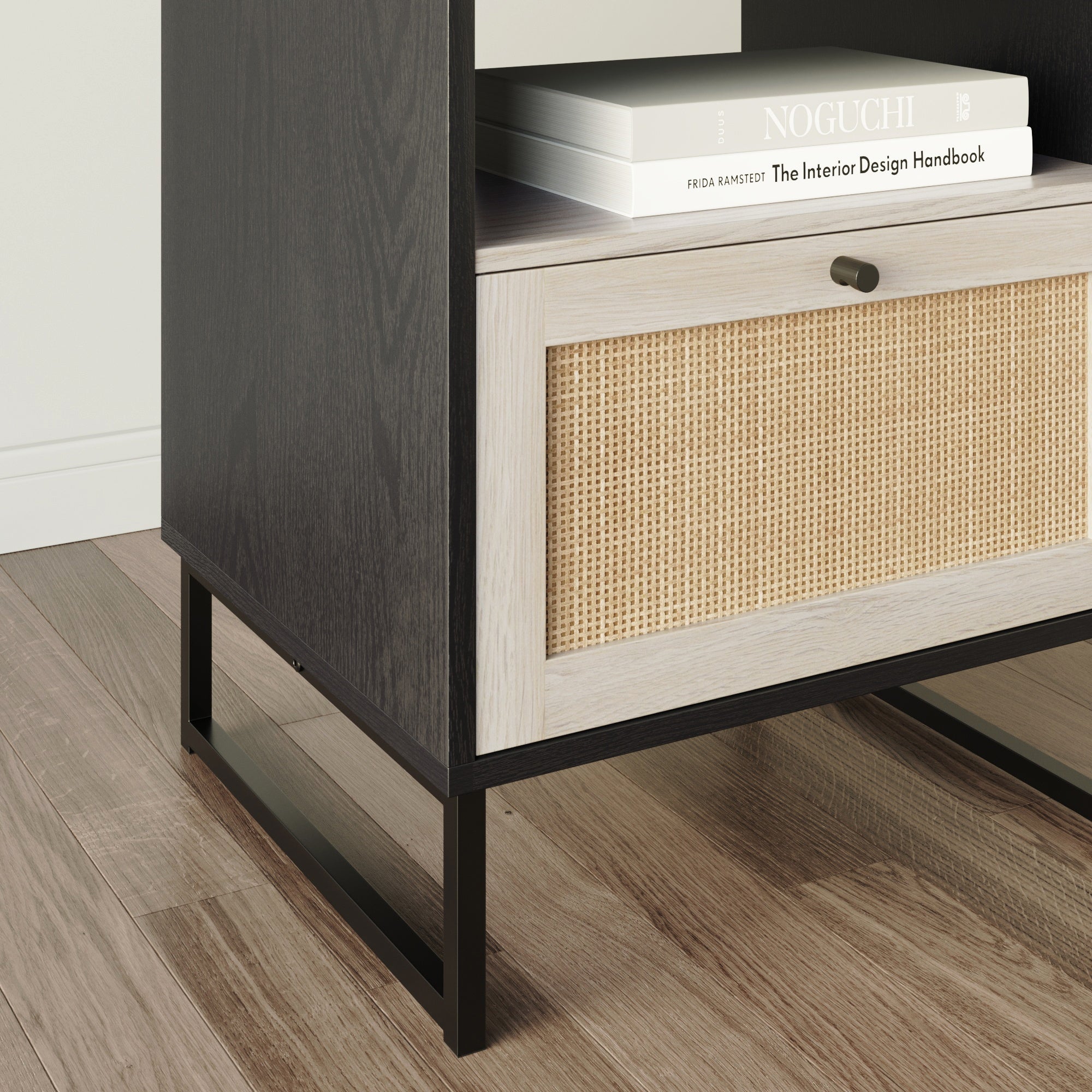A close-up of a modern cabinet with black metal legs, a woven rattan drawer with a round knob, and a book titled The Interior Design Handbook on top, on a wooden floor.