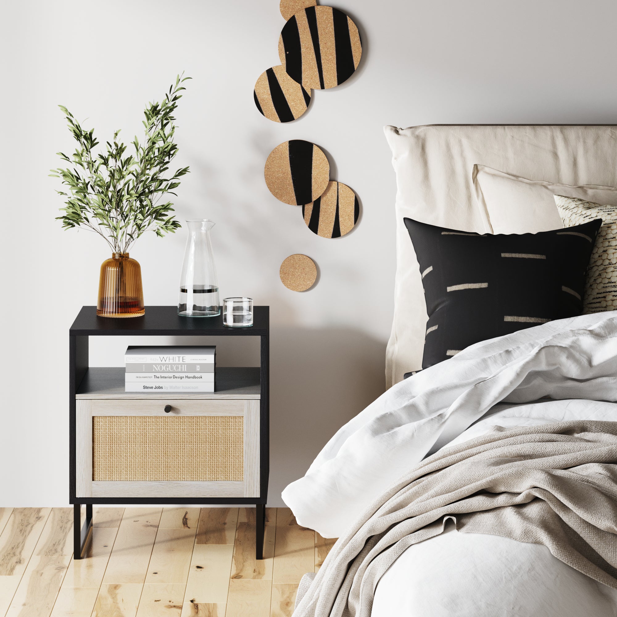 A modern bedroom features a bed with neutral bedding, a black and beige pillow, a nightstand with books, a glass carafe, an amber vase with leafy branches, and abstract circular wall art above light wood floors.