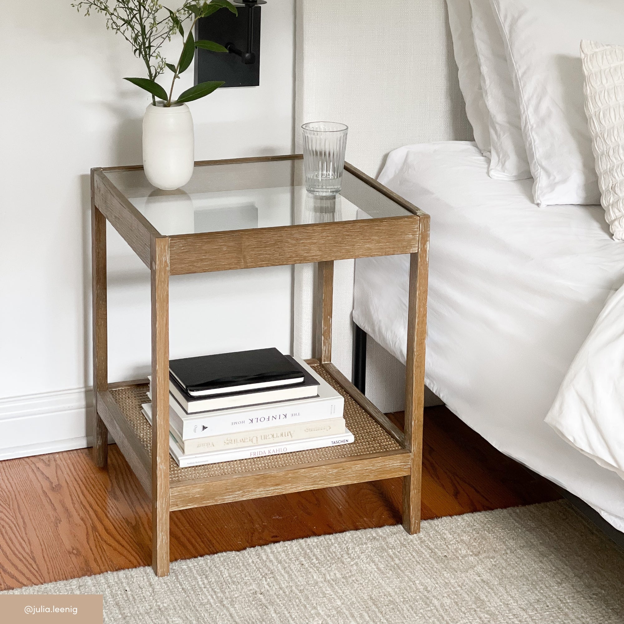 A wooden bedside table with a glass top holds a white vase with greenery and a clear glass of water. Below, several stacked books rest on the lower shelf. The table is next to a white bed on a wooden floor.
