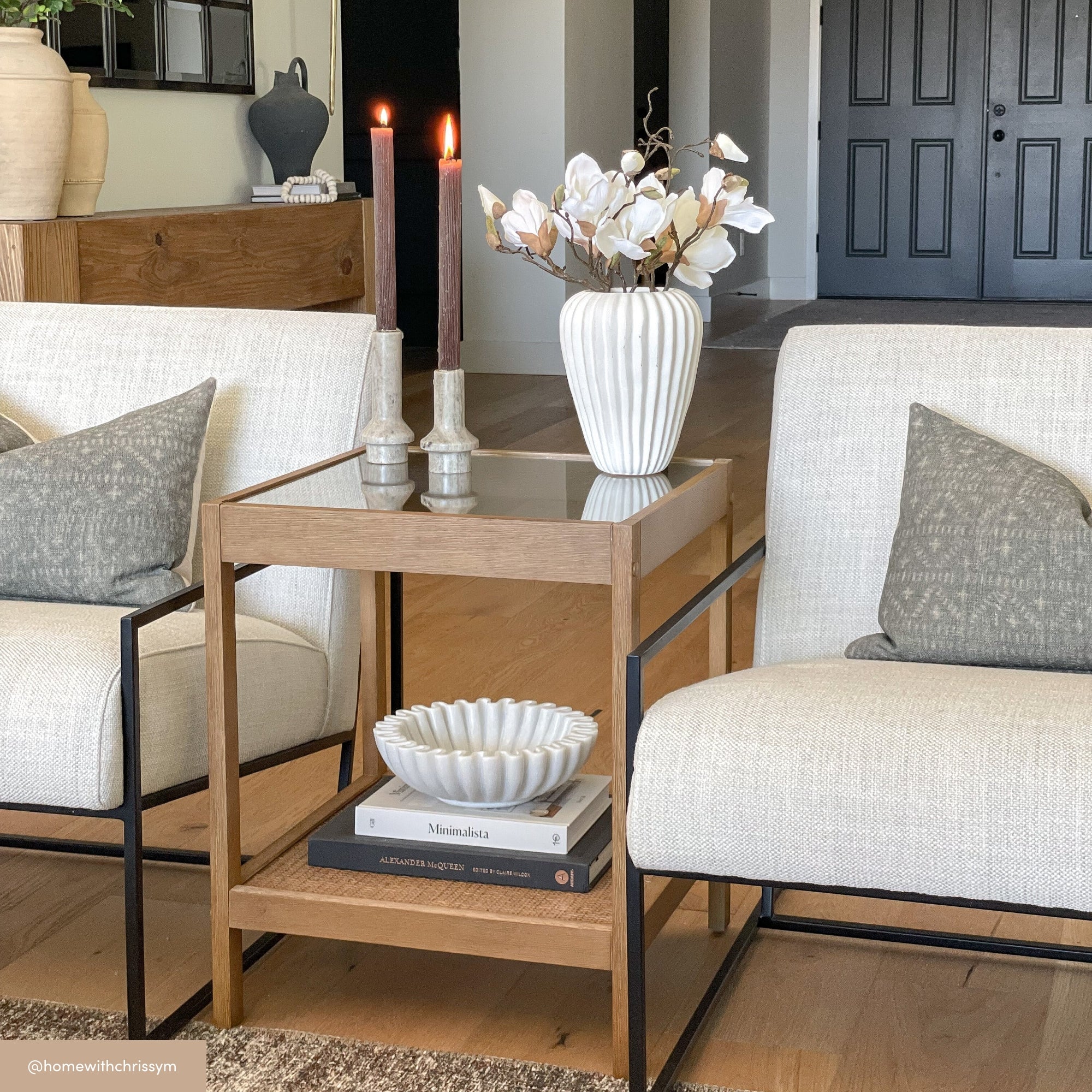 A modern living room featuring two cream armchairs, a wooden side table with a glass top, two lit tall candles, a white vase with white flowers, and stacked books, set against a neutral background with double doors.