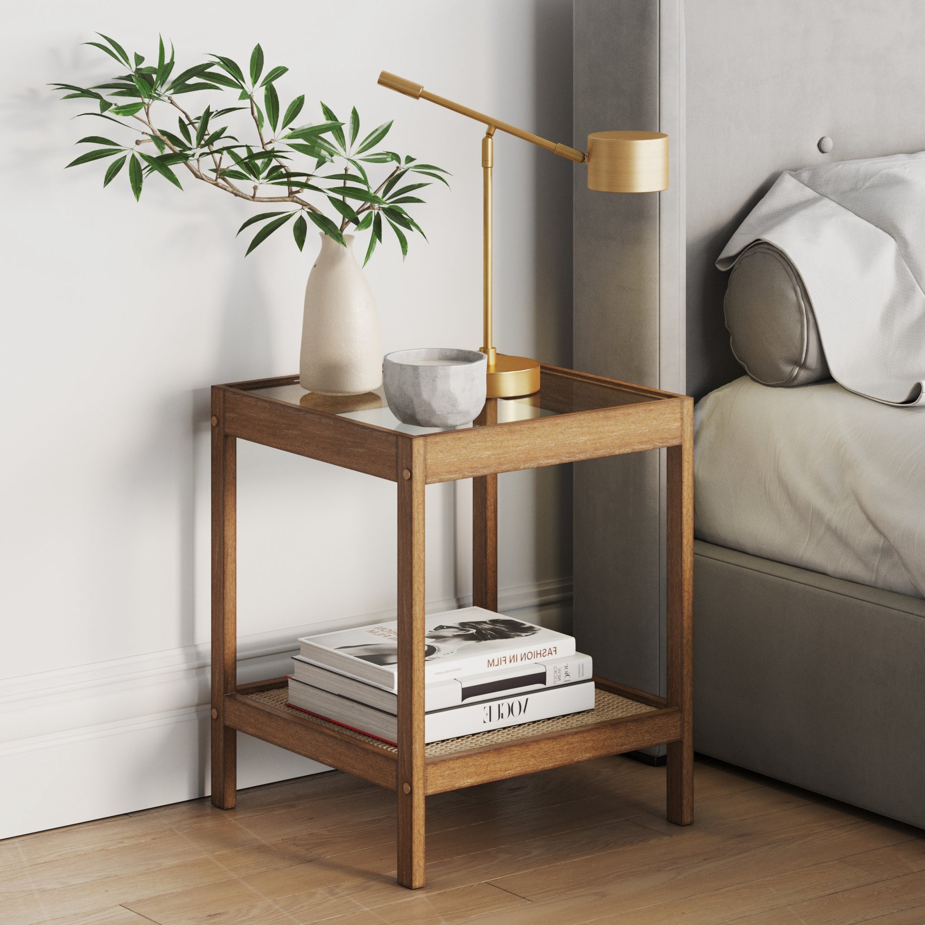 A wooden bedside table with a potted plant, a gold desk lamp, a small bowl, and books on the lower shelf stands next to a gray upholstered bed on a wooden floor.