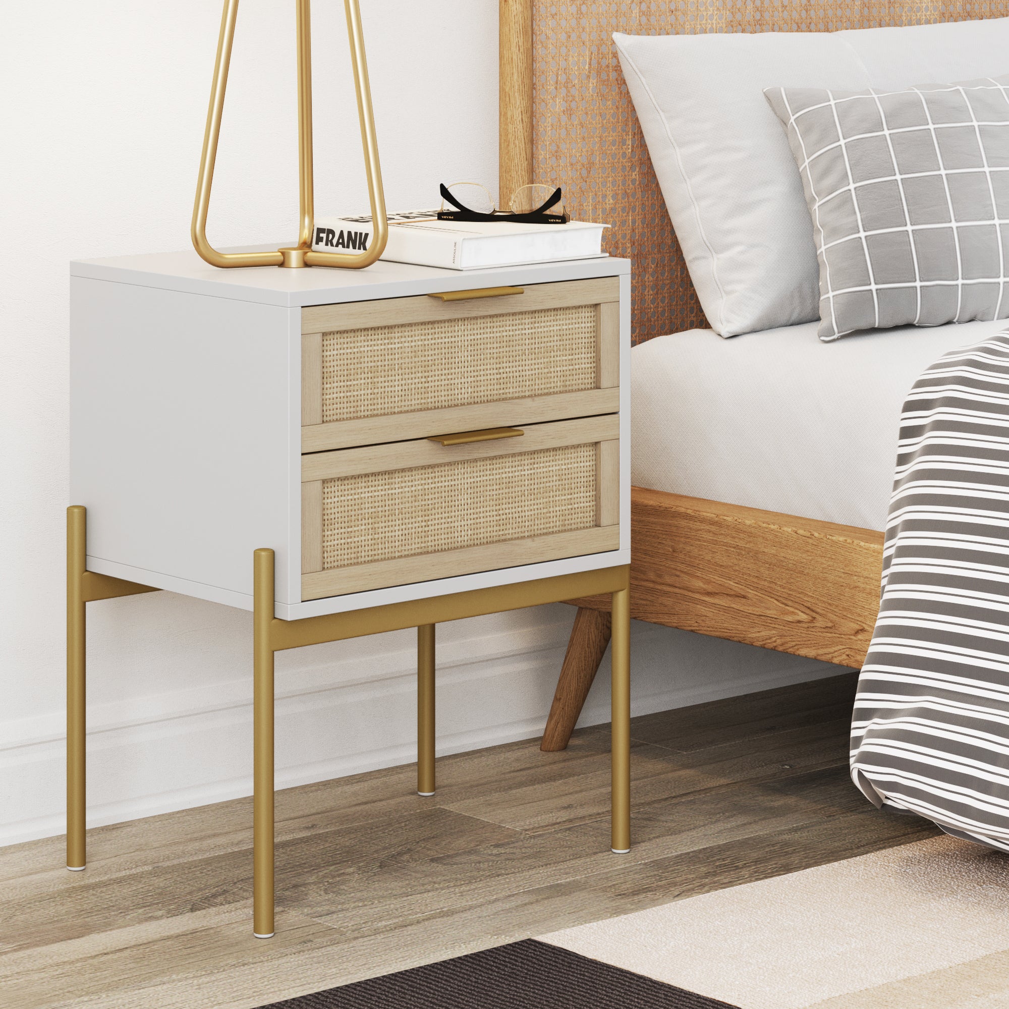 A modern nightstand with two wood and rattan drawers, gold legs, and a white frame sits beside a bed with gray plaid bedding. The nightstand holds a lamp, glasses, and a small book.