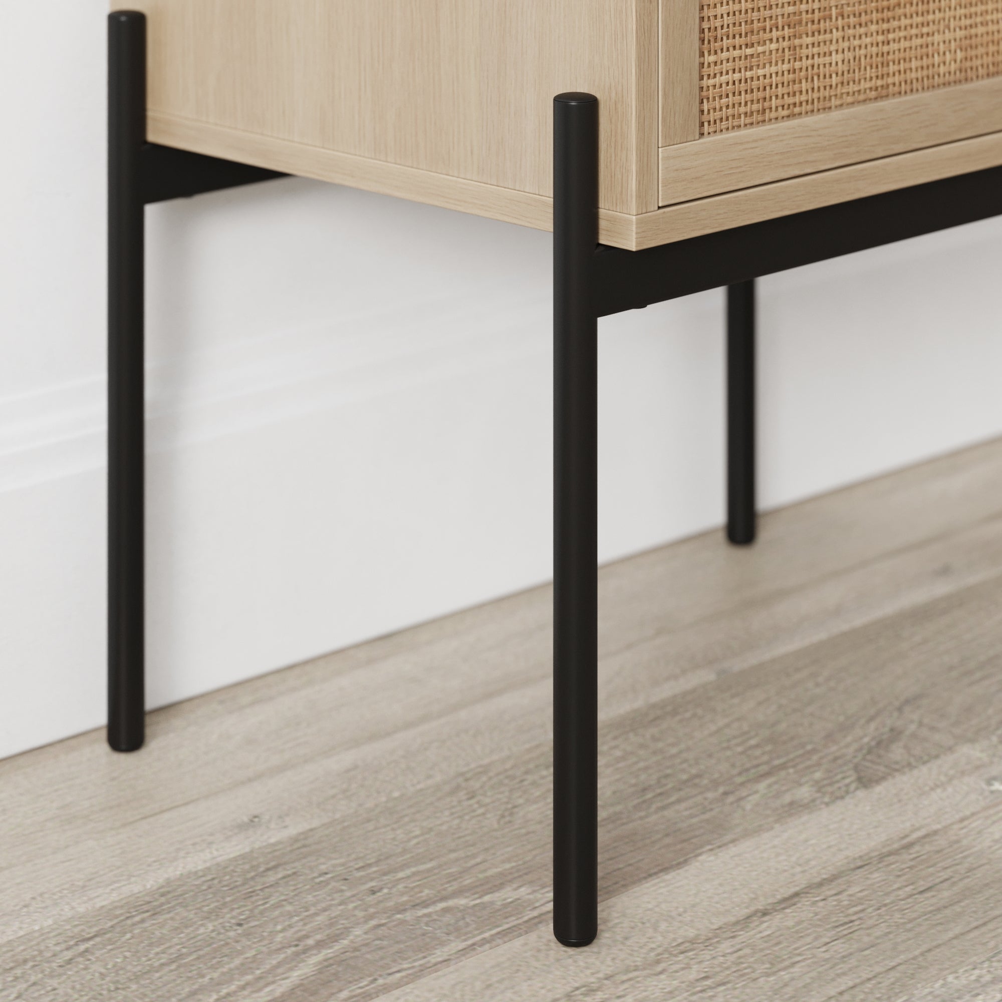 A close-up of the black metal legs of a light wood cabinet with a woven rattan panel, standing on light-colored wood flooring against a white wall.