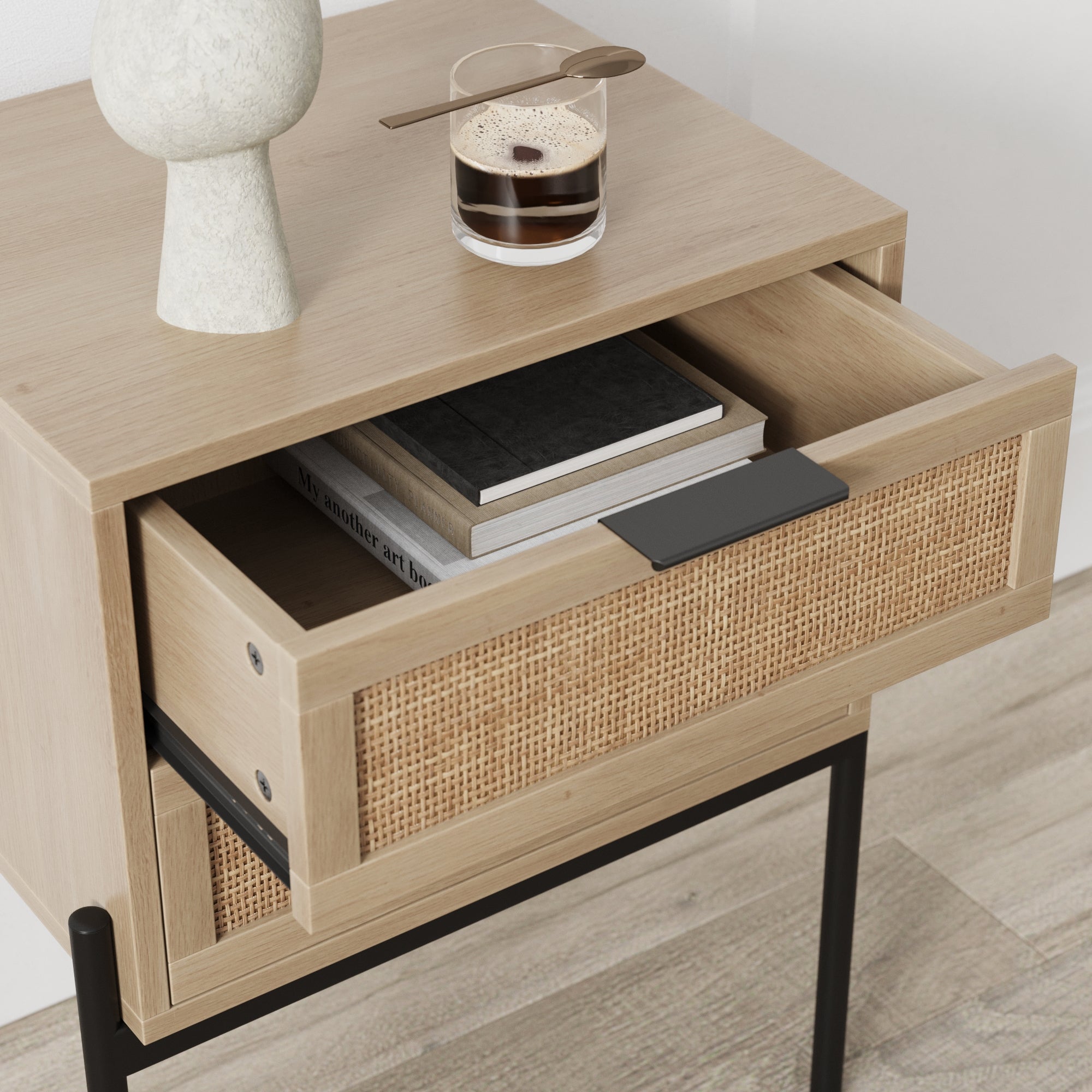 A light wood nightstand with a woven front drawer, partially open to reveal stacked books, a decorative ceramic object on top, and a glass of coffee with a spoon resting on the rim.