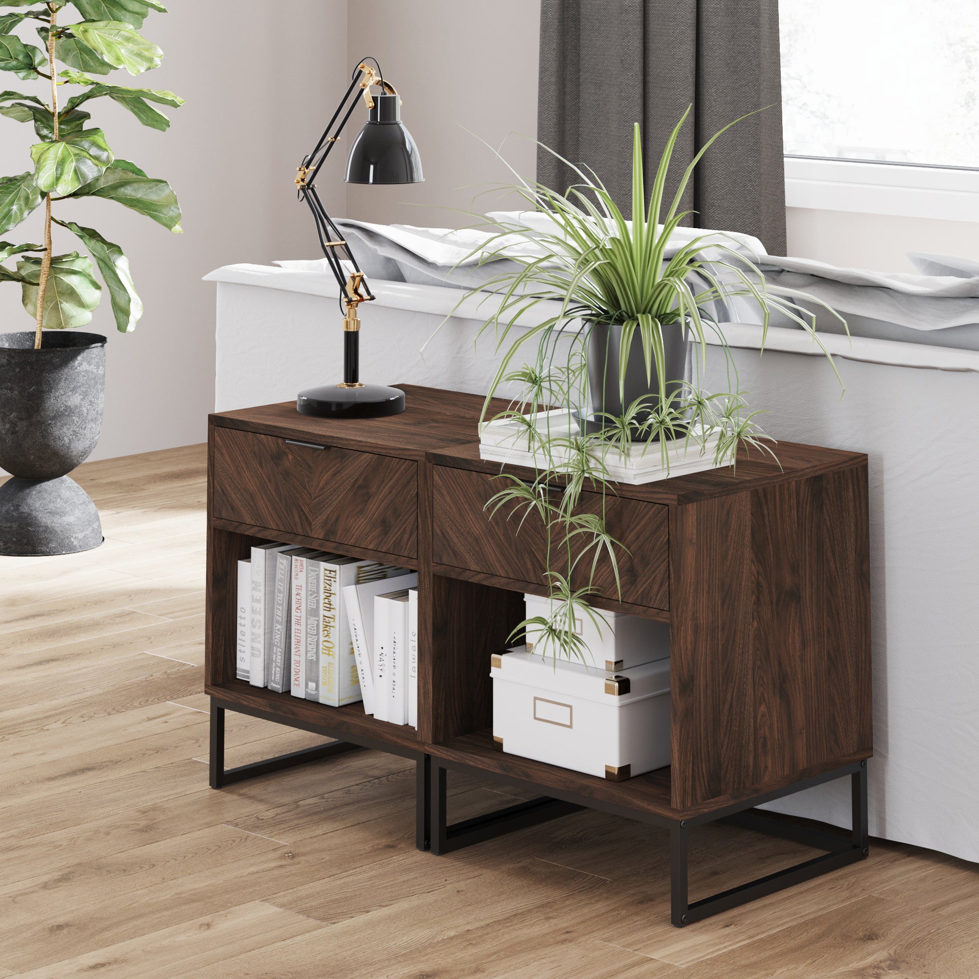 A modern wooden console table with books, a potted plant, storage boxes, and a black desk lamp sits behind a sofa in a bright room with wood floors, a large window, and a tall leafy plant in a black pot.