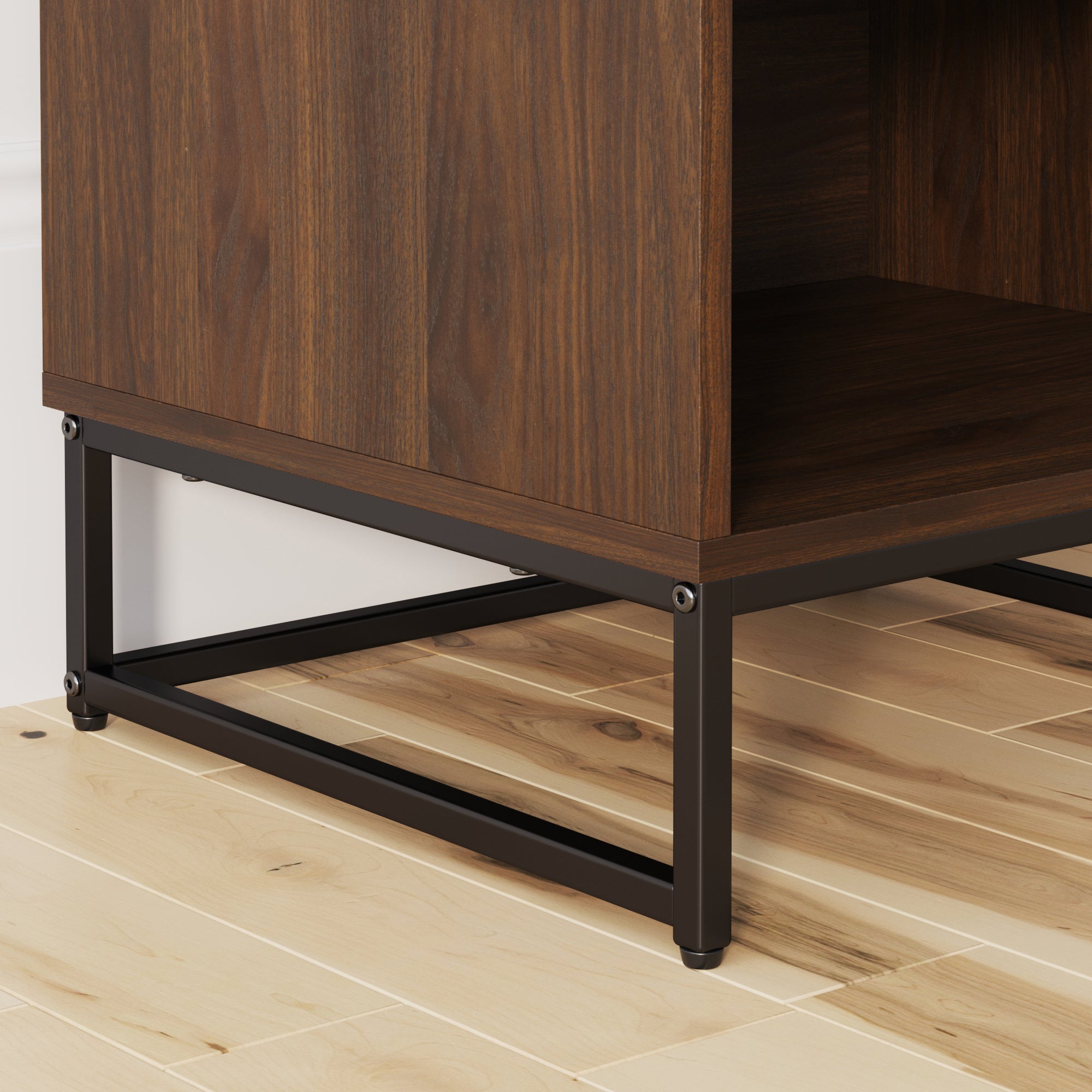 Close-up of the bottom corner of a wooden cabinet with a dark metal frame, resting on a light-colored hardwood floor near a white wall.