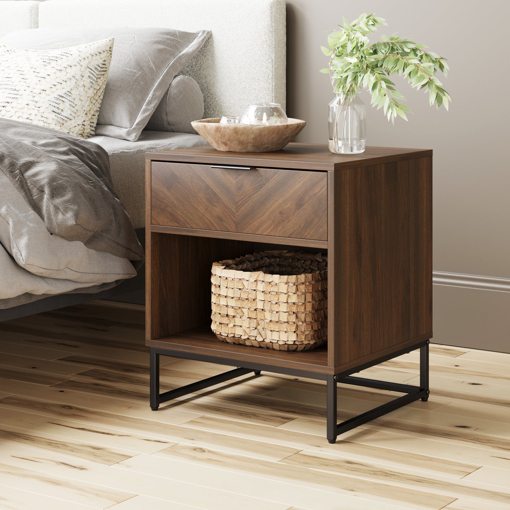 A modern wooden nightstand with a drawer and open shelf, featuring a woven basket inside. On top, there is a small tray, a glass vase with green leaves, and a glass. Beside it, a bed with gray bedding and pillows.