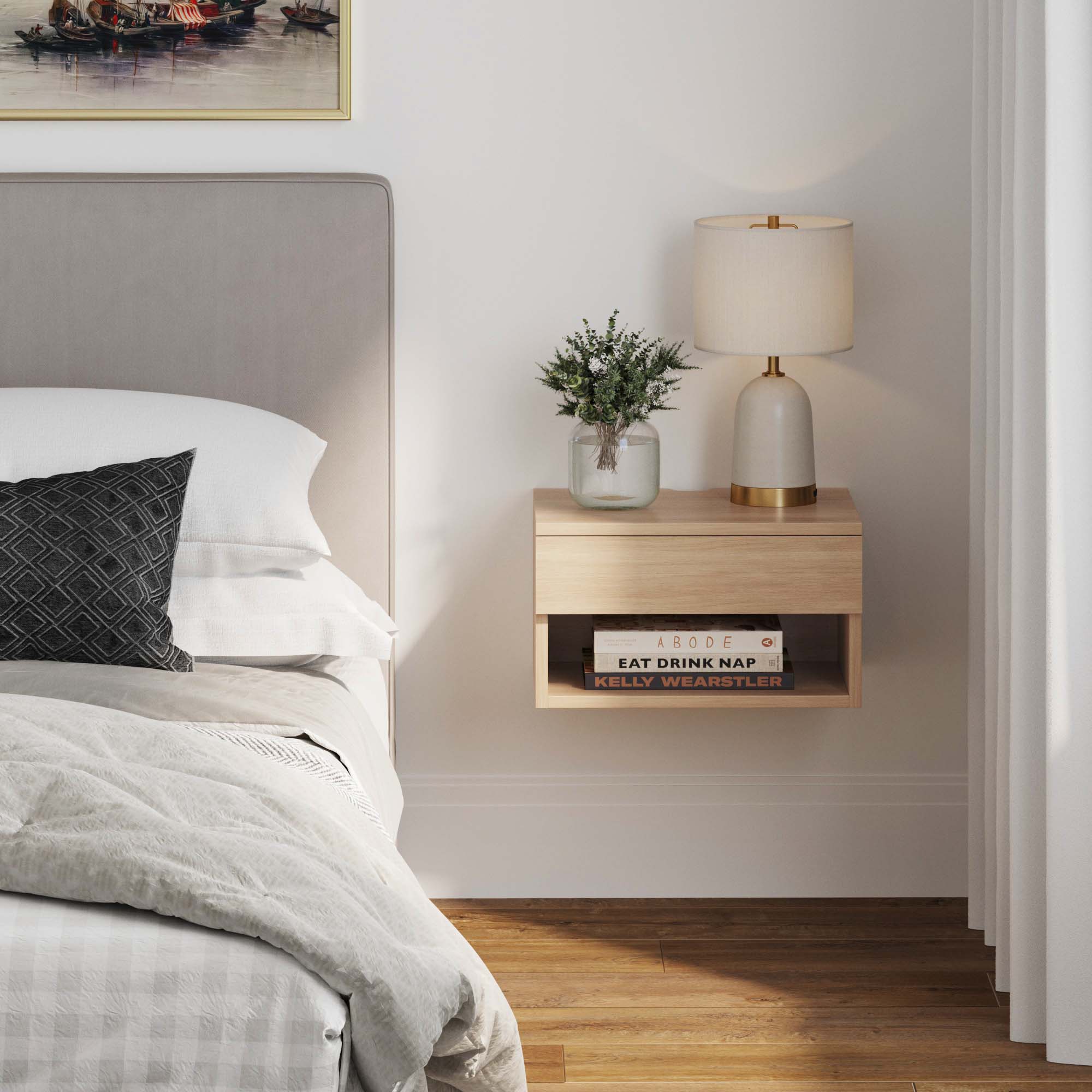 A modern bedroom with a floating wooden nightstand holding a lamp, a glass vase with greenery, and books. The bed has white linens, a gray headboard, and a black pillow. Sunlight filters through white curtains.