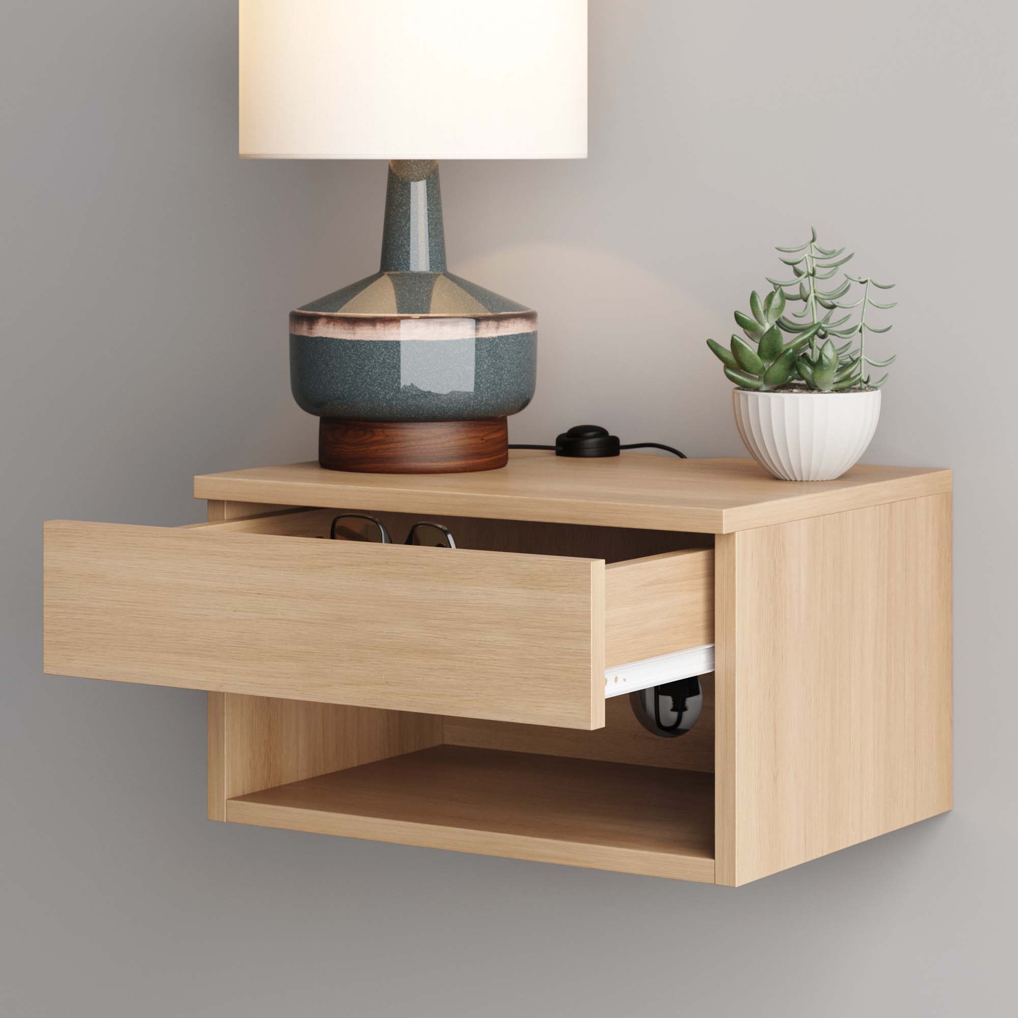 A light wood floating nightstand with an open drawer, revealing a pair of glasses inside. On top, there is a lamp with a ceramic base and a small potted succulent. A white wall is visible in the background.