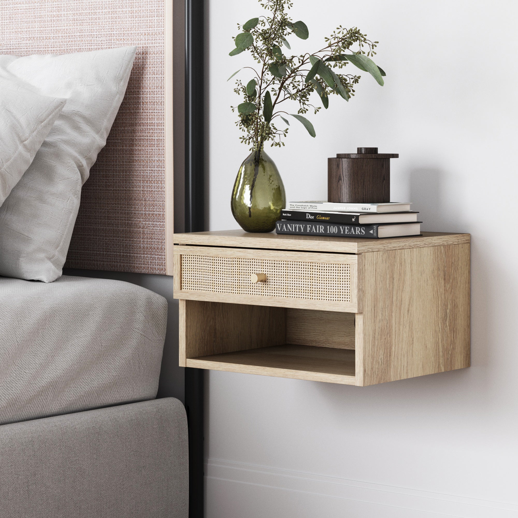 A light wood floating nightstand with a rattan-front drawer, open shelf, and decorative items on top, including a green vase with foliage, stacked books, and a small wooden container, next to a bed.
