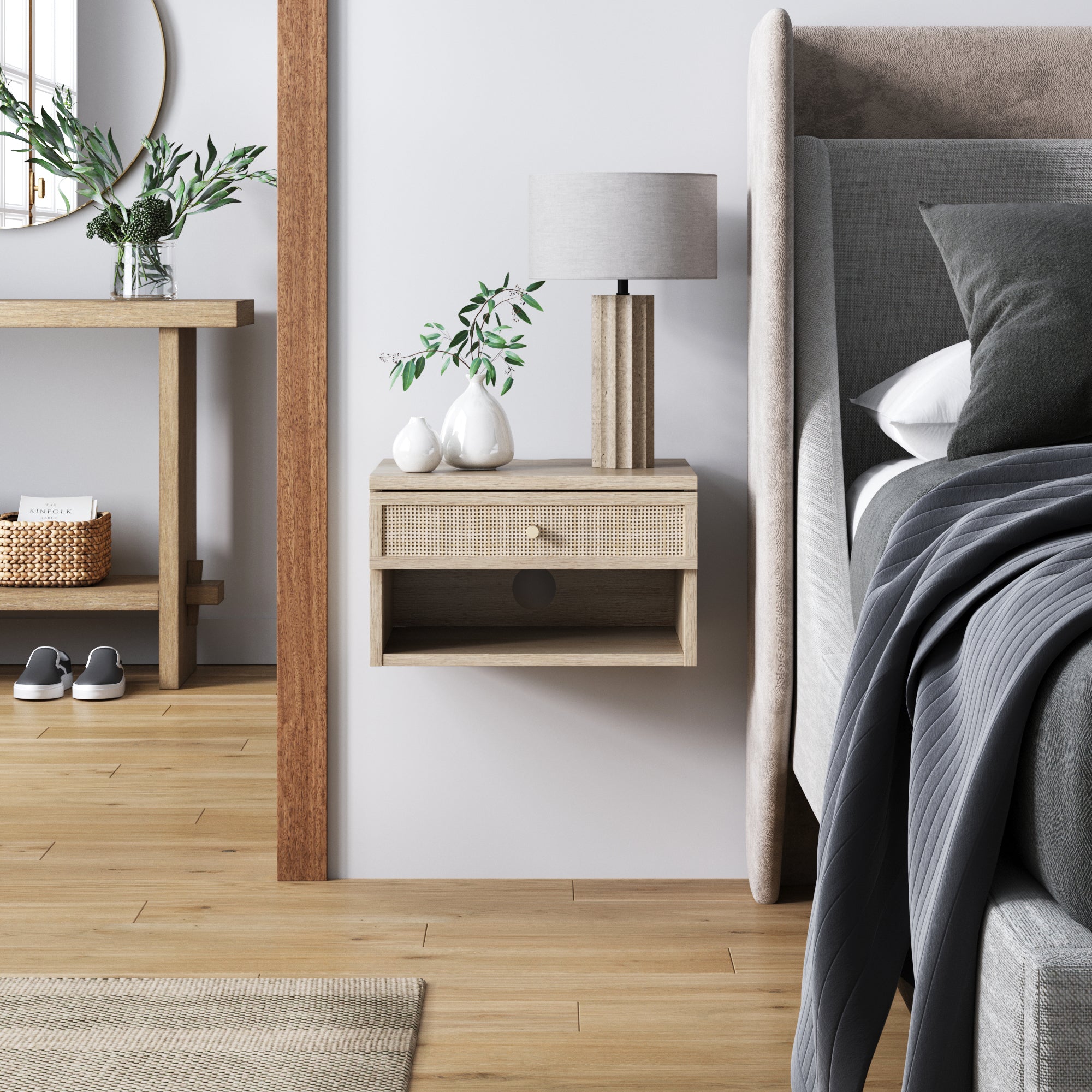 A modern bedroom features a floating wooden nightstand with a drawer, a gray lamp, and decorative ceramic vases. The bed has gray bedding, and a console table with plants and shoes is seen in the background.