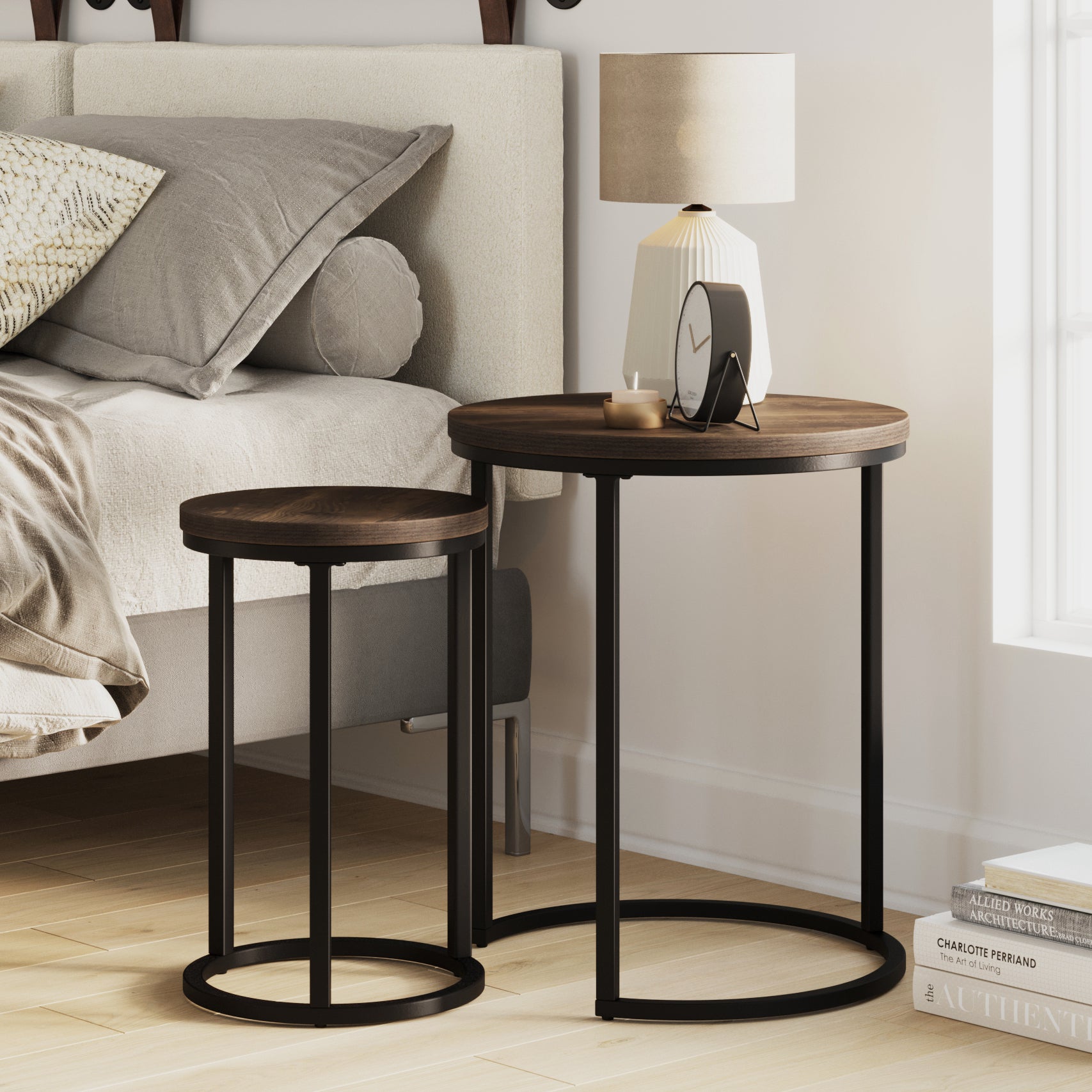 Two round wooden nesting tables with black metal frames beside a bed. The larger table holds a lamp, a small clock, and a cup. Sunlight streams through a nearby window onto the light wooden floor.