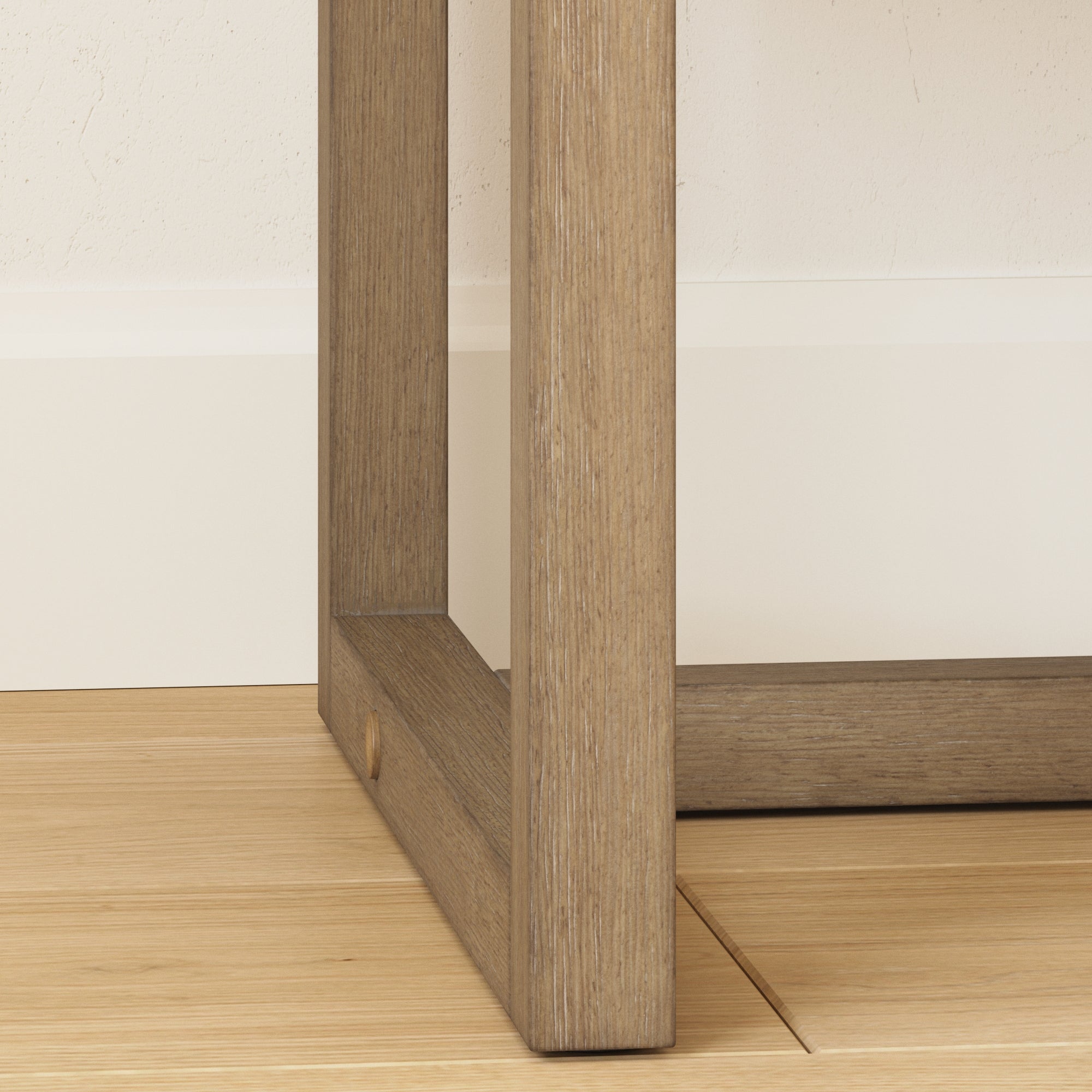 Close-up of the bottom corner of a wooden table or furniture leg on a light wood floor, next to a white baseboard and textured off-white wall.