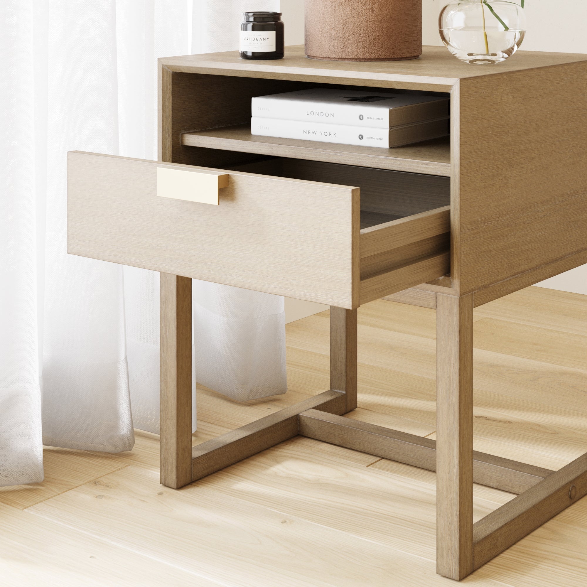 A light wood nightstand with an open drawer, books and small decorative items on top. Sunlight filters through a sheer white curtain onto the light wooden floor.