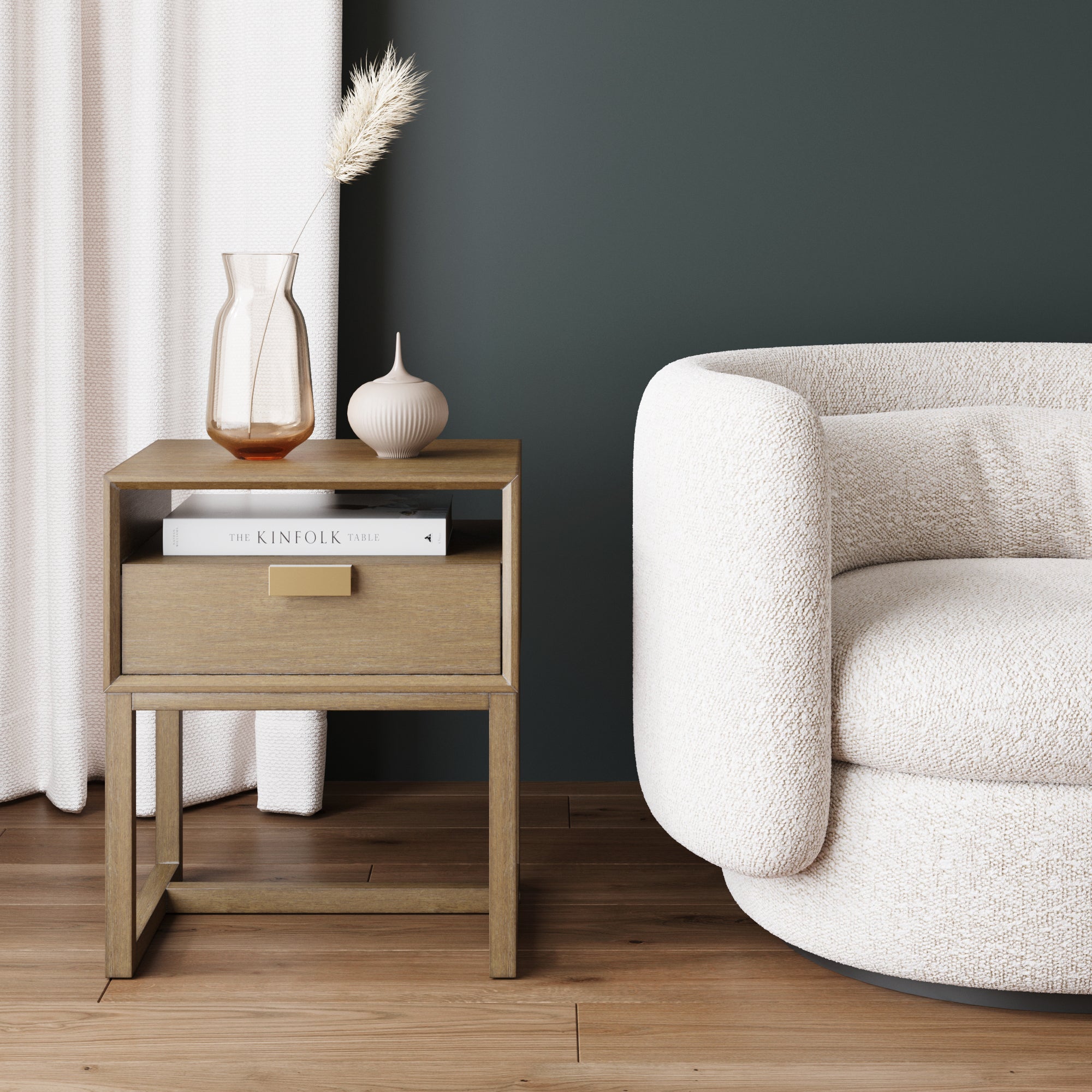 A modern wooden side table with a drawer holds two books and two vases, one with dried grass. Next to it is a white textured armchair, set against a dark wall and light curtains on a wooden floor.