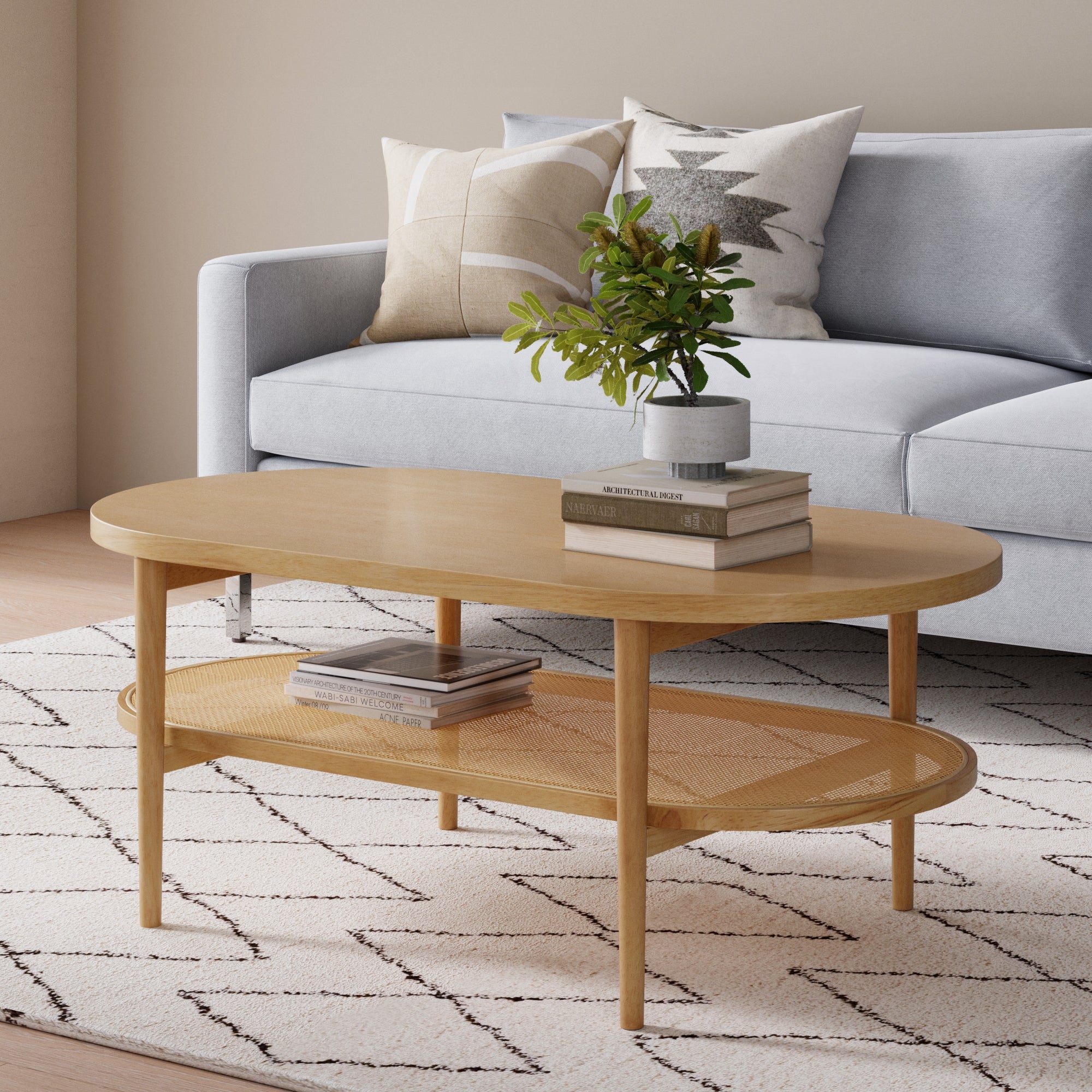 A modern living room with a light gray sofa, patterned pillows, and a wooden oval coffee table holding books and a vase with green leafy branches, set on a white rug with a geometric design.