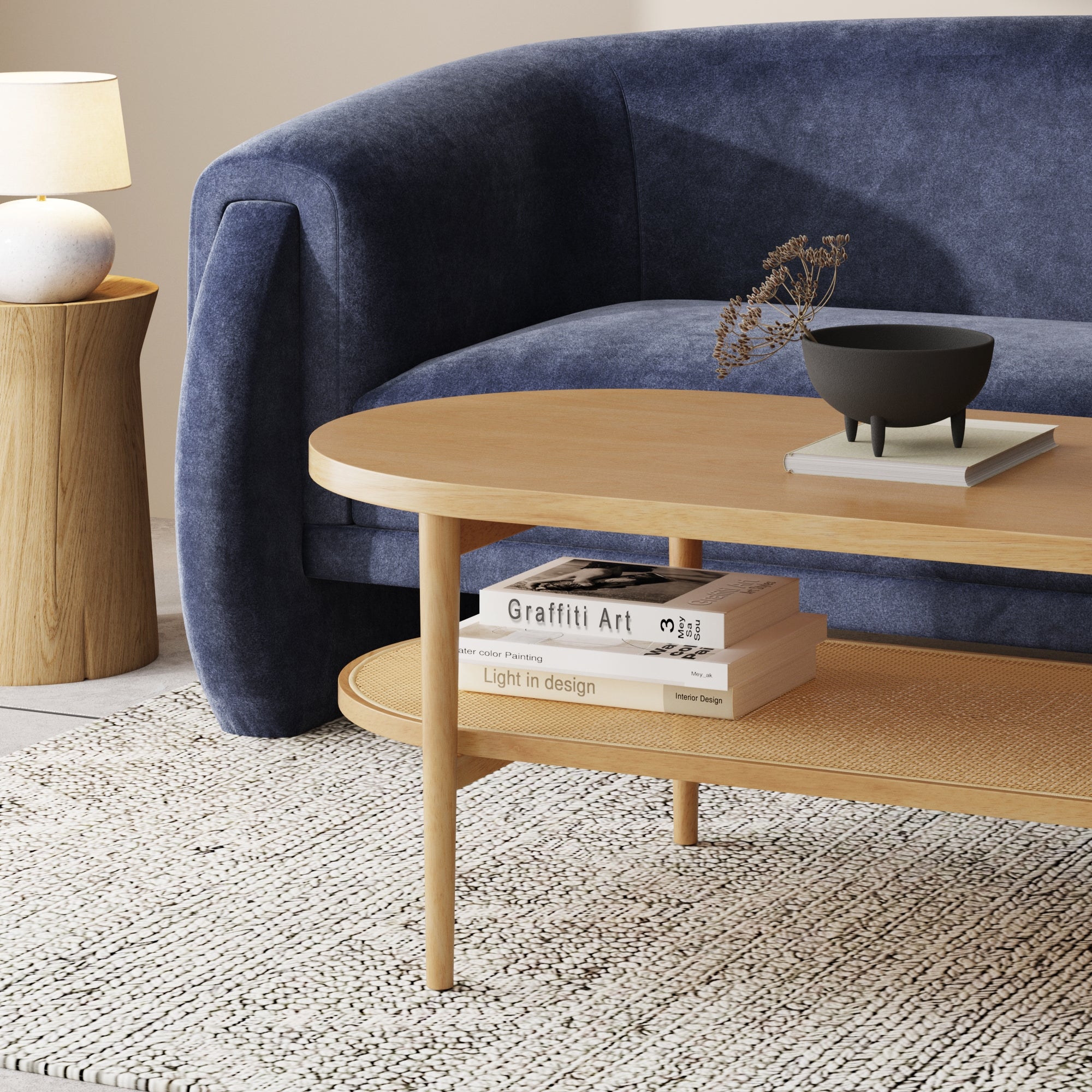 A modern living room with a blue velvet sofa, a wooden coffee table holding books and a decorative bowl, a round wooden side table with a lamp, and a patterned rug on the floor.