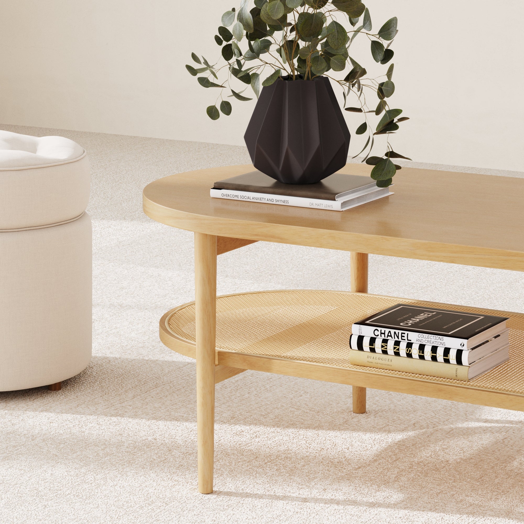 A wooden coffee table with a black geometric vase holding green foliage and two books on top. A Chanel book sits on the lower shelf. The table is set on a light carpet next to a round, white cushioned seat.