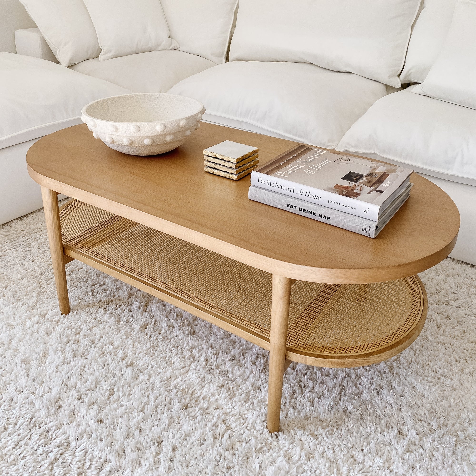 A light wood oval coffee table with a lower woven shelf holds a textured white bowl, coasters, and stacked books, all set on a white textured rug in front of a white sofa.