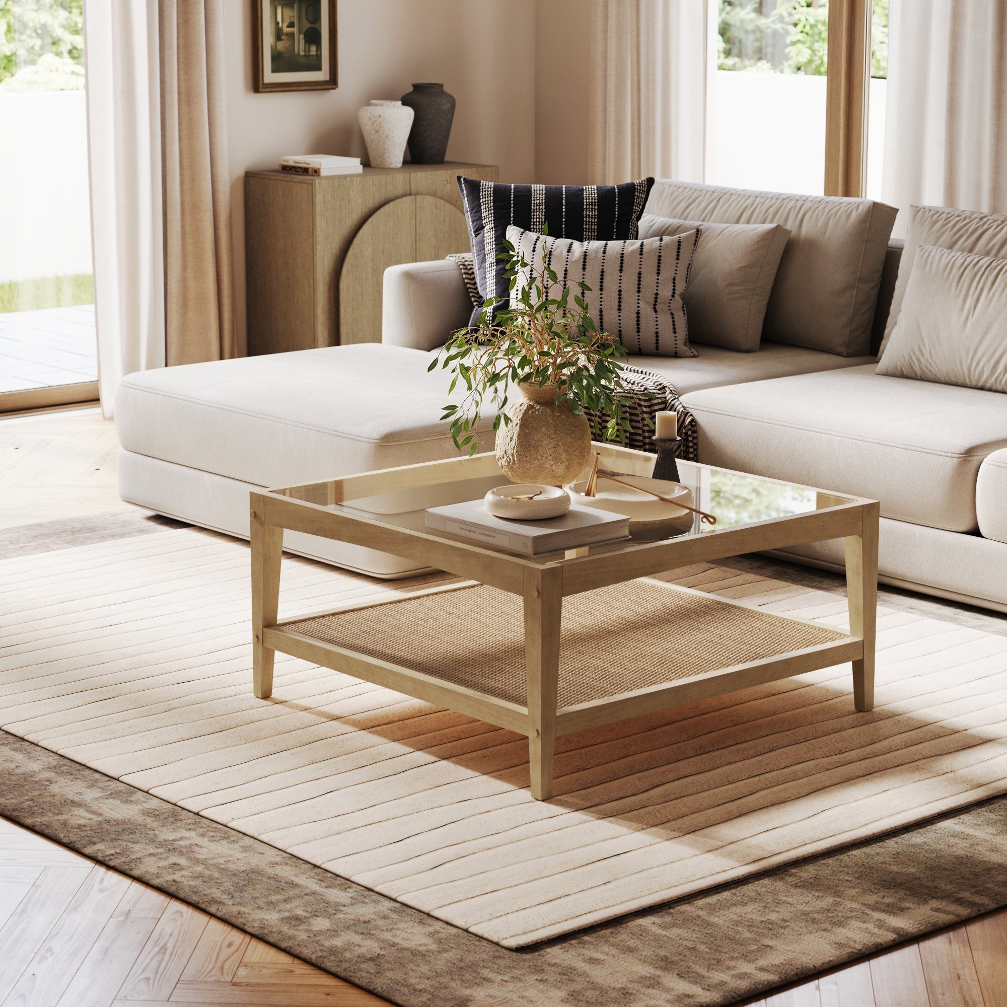 A modern living room with a beige sectional, textured pillows, and a Nathan James Square Glass Coffee Table with solid wood legs and rattan shelf, topped with greenery, on neutral rugs near large windows with light curtains.