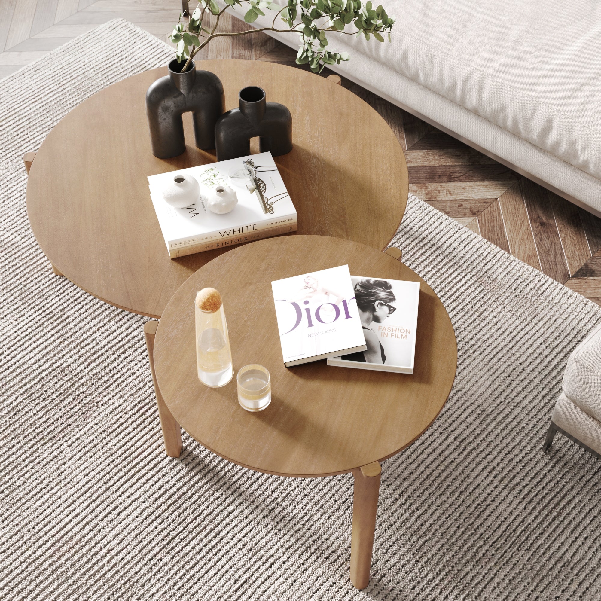 Two round wooden coffee tables overlap on a textured rug. On the tables are black vases, books, a clear bottle, and glasses of water. A beige sofa and wood herringbone floor are partially visible.