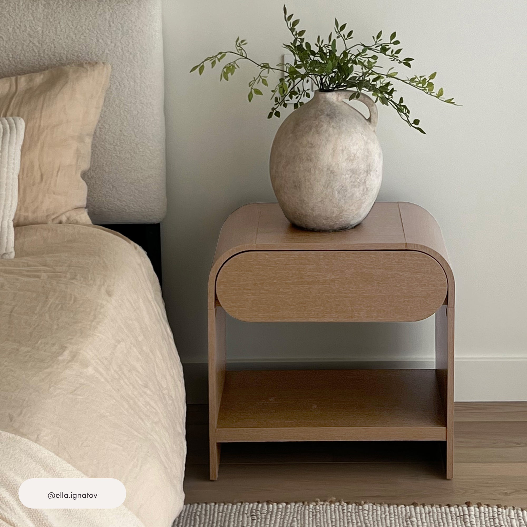 A minimalist wooden nightstand beside a bed with beige bedding. On the nightstand is a large, rustic ceramic vase holding green leafy branches. The scene is set against a plain, light-colored wall.