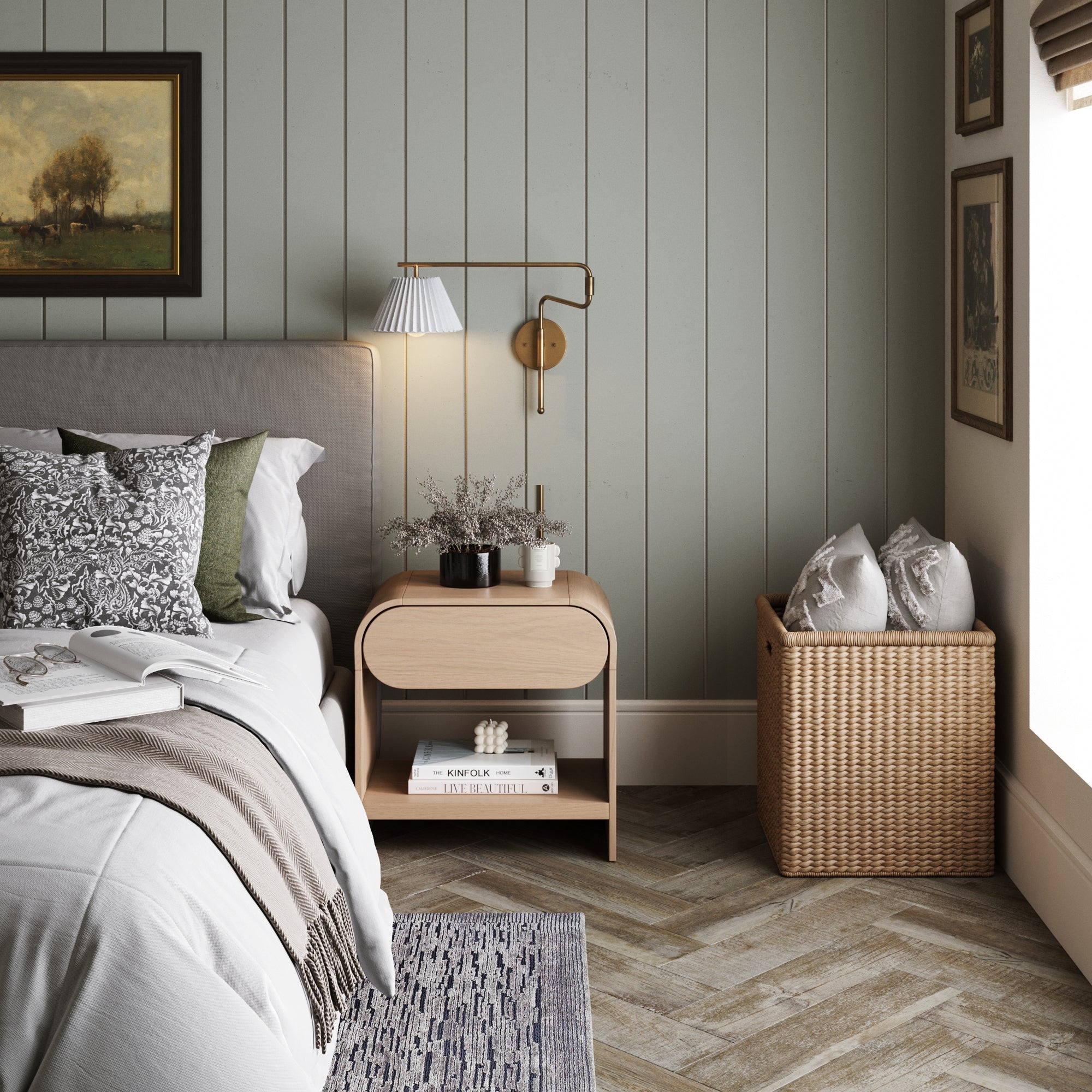 A cozy bedroom corner with a bed, patterned pillows, a wooden nightstand with a lamp and vase, framed art on pale paneled walls, and a wicker basket filled with pillows on a herringbone wood floor.