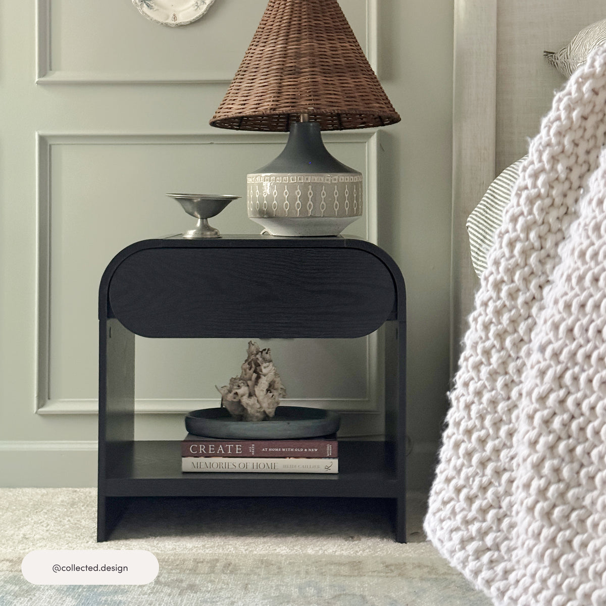 Black Mid-Century Wood Arched Nightstand in black oak with an open shelf holding books and a decorative object, topped with a small silver bowl and a woven lamp.