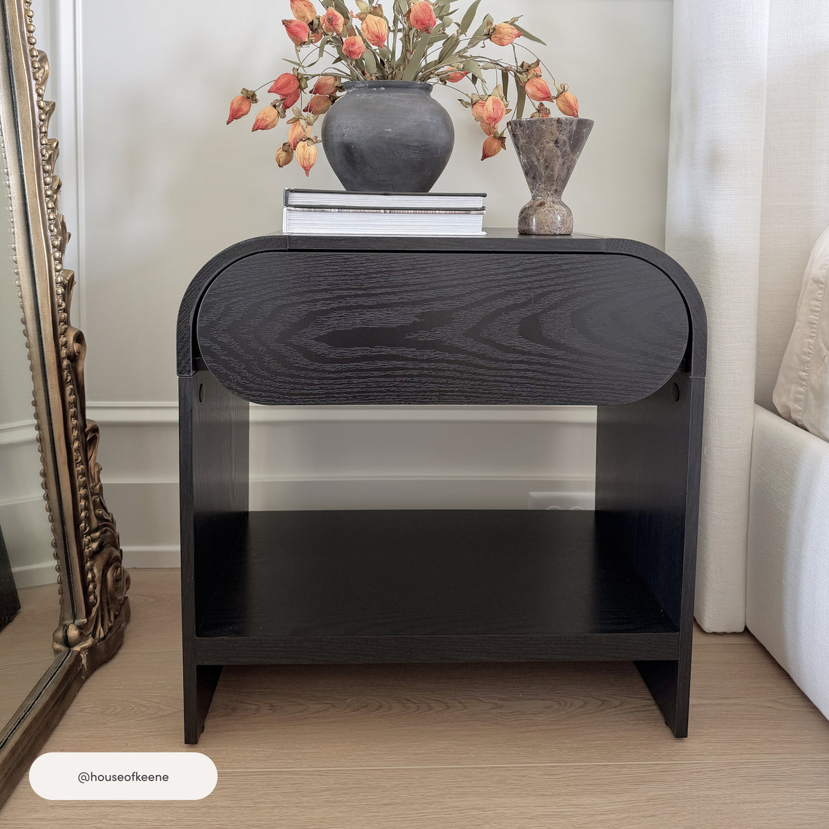 Nathan James Mid-Century Wood Arched Nightstand Black Oak with an open shelf and a top drawer, topped with a vase of dried flowers and stacked books.