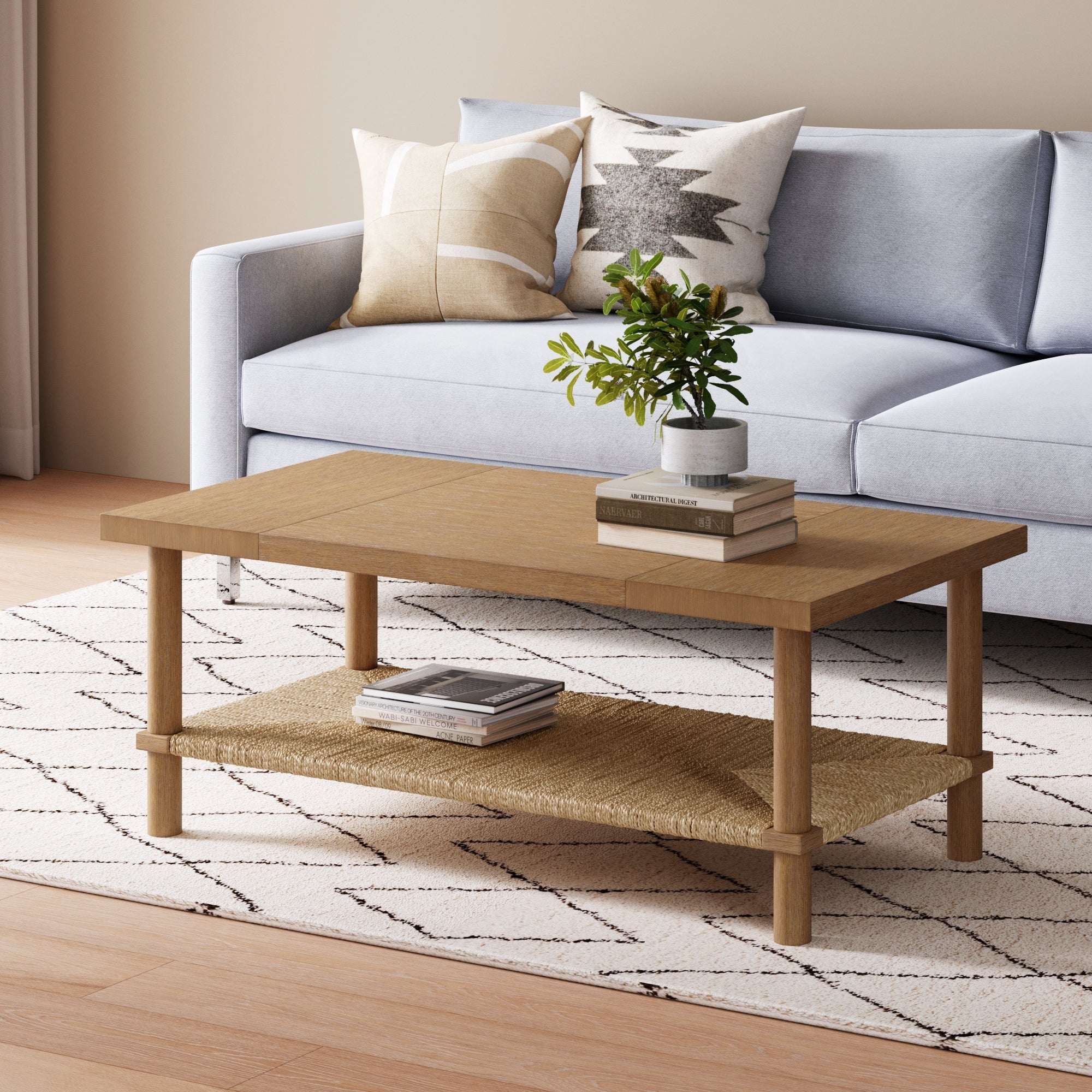 A light wood coffee table with books and a small plant sits on a patterned rug in front of a light blue sofa with decorative cushions in a cozy living room.