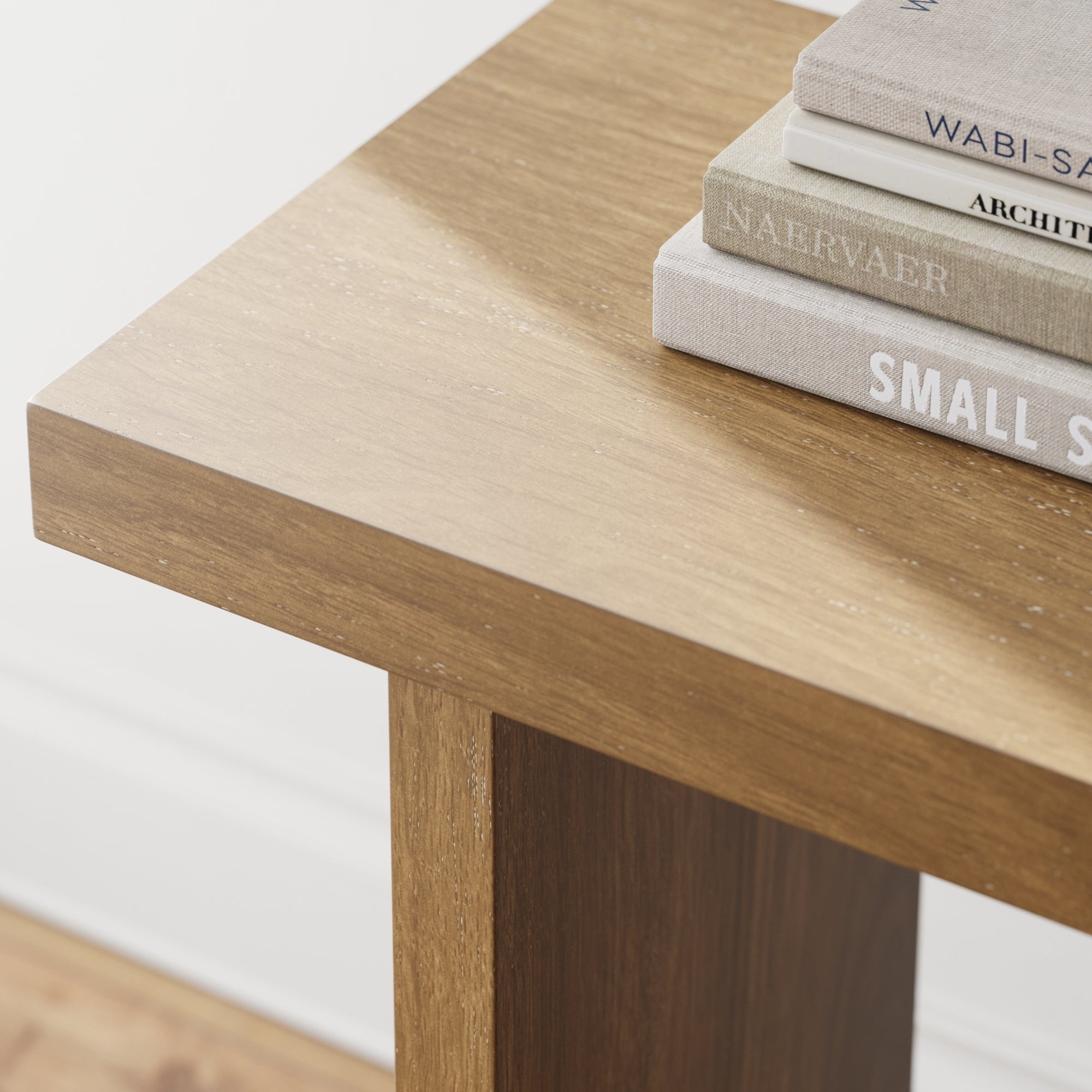 A close-up of a wooden table corner with a stack of books on top, showing the table’s natural wood grain and a clean, minimalist design. The edge of the table and part of the white wall and floor are visible.