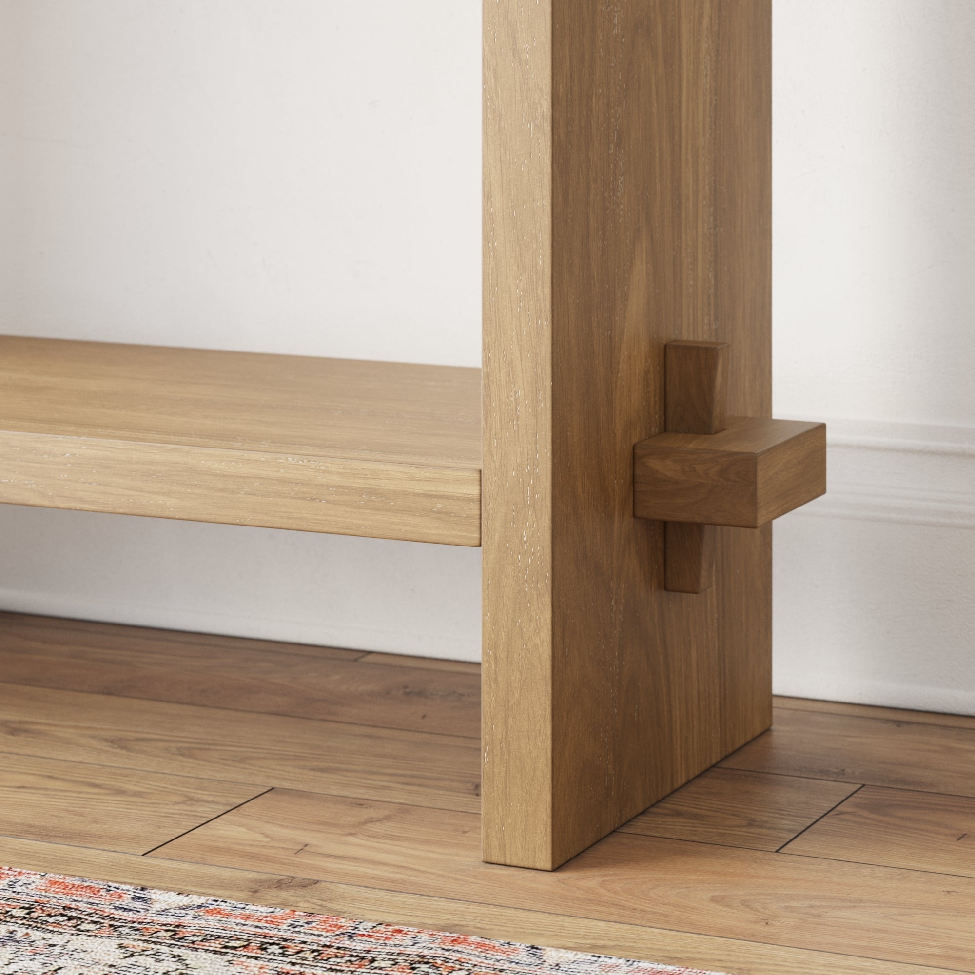 A close-up of a wooden shelf unit with a unique joint detail, showing the vertical support intersected by a perpendicular wooden block. The shelf stands on a wooden floor next to a white wall and a patterned rug.