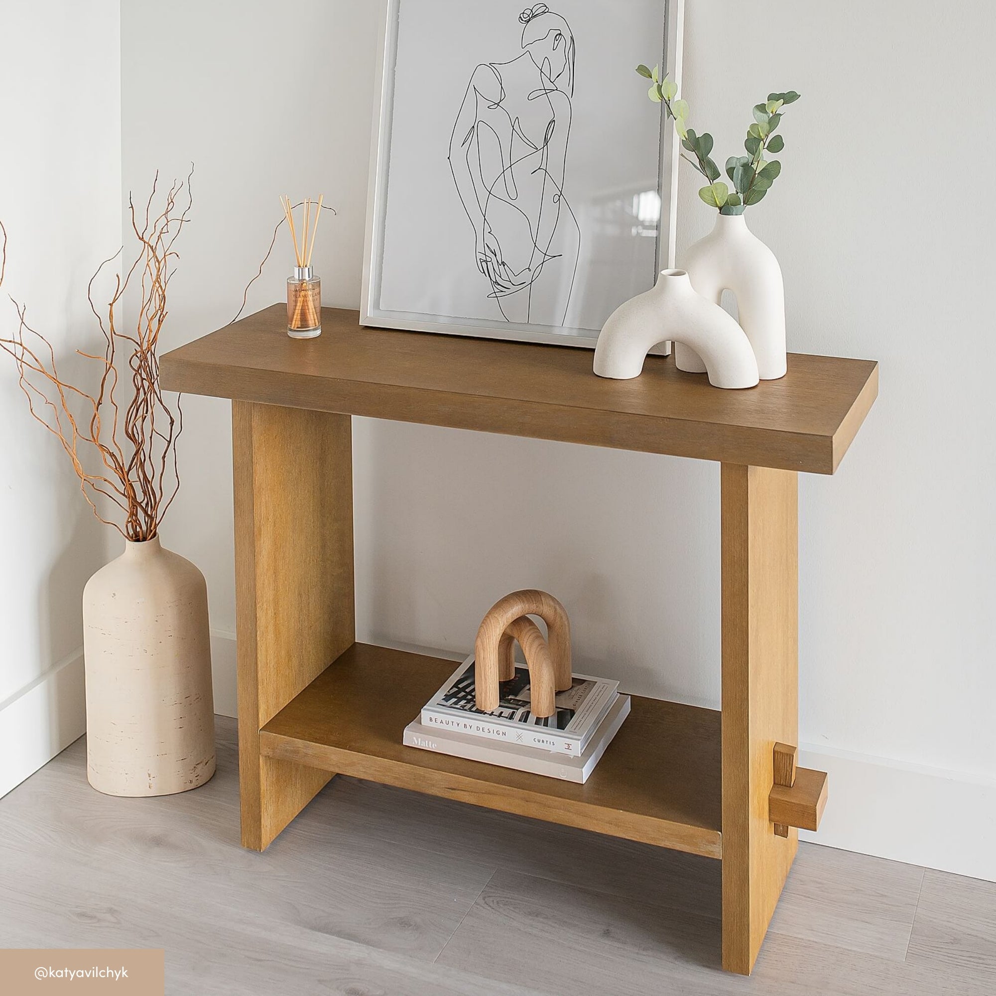 A wooden console table against a light wall features art, a white vase with greenery, a reed diffuser, decorative branches in a floor vase, and books with a curved wooden sculpture on a lower shelf.