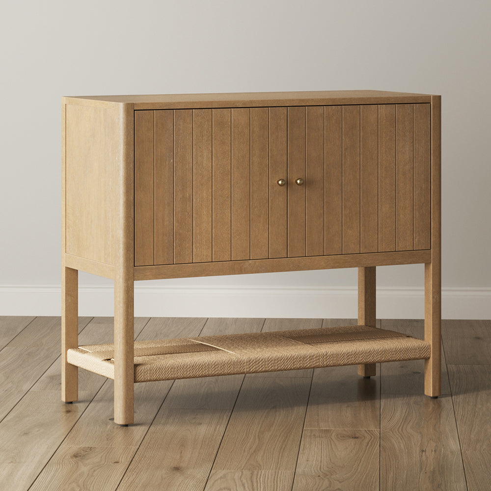 Light wood cabinet with two fluted doors and a woven lower shelf.