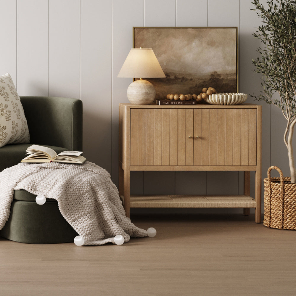 A wooden cabinet with a lamp, landscape painting, and decorative bowl sits next to an upholstered green chair draped with a chunky knit blanket.