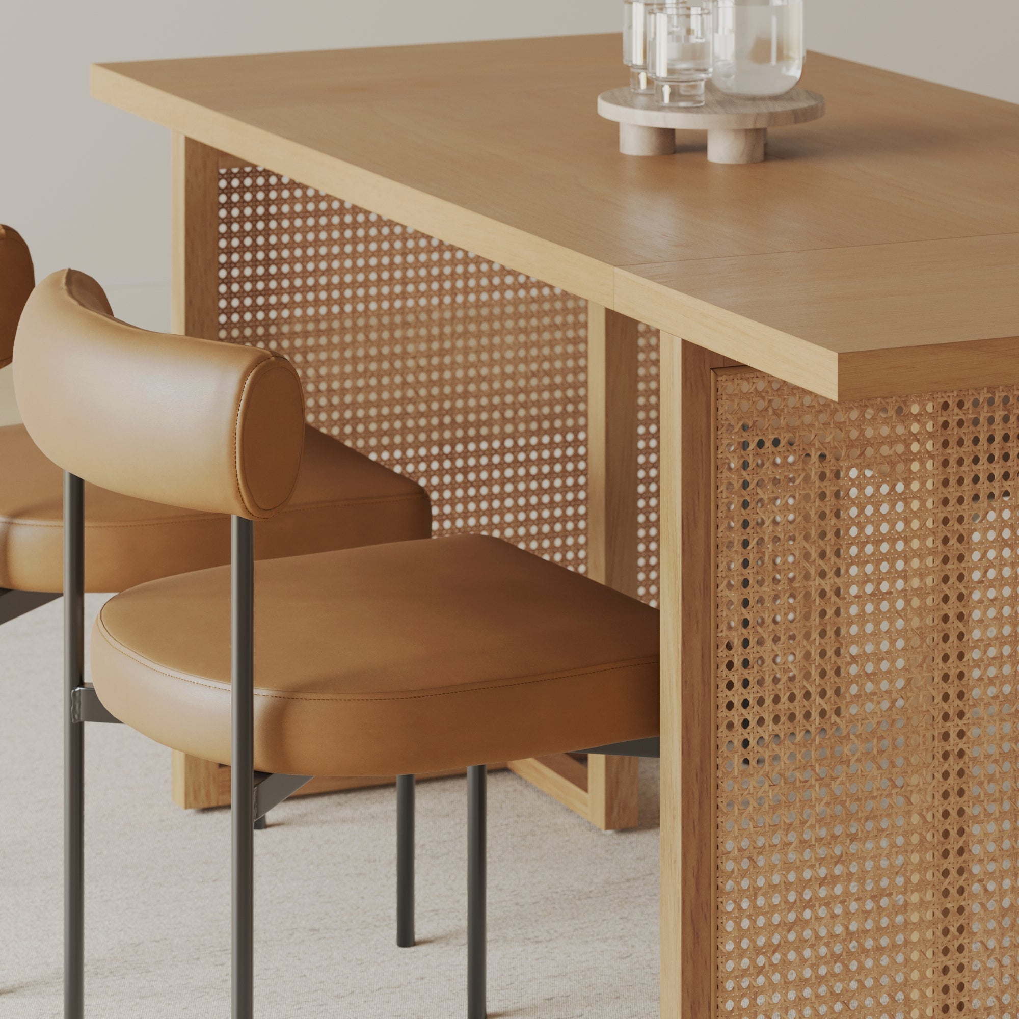 A wooden table with a woven cane panel on the side is paired with two tan leather chairs featuring rounded backs and metal legs, set on a light-colored carpet. Two glass candles sit on the table.