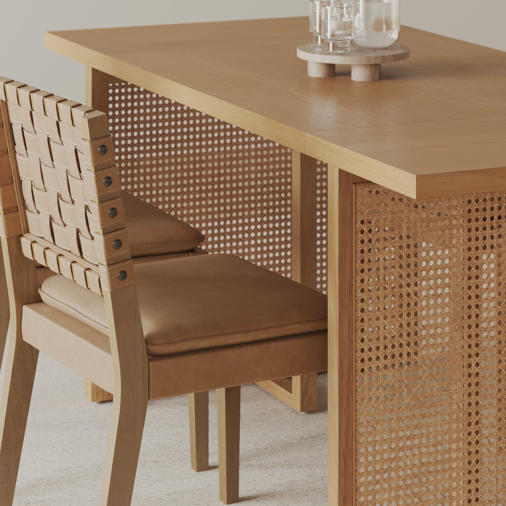 A light wood table with a woven cane panel on the side is paired with a tan leather chair featuring a woven backrest. Glassware sits on a tray atop the table, and the setting is on a light-colored carpet.