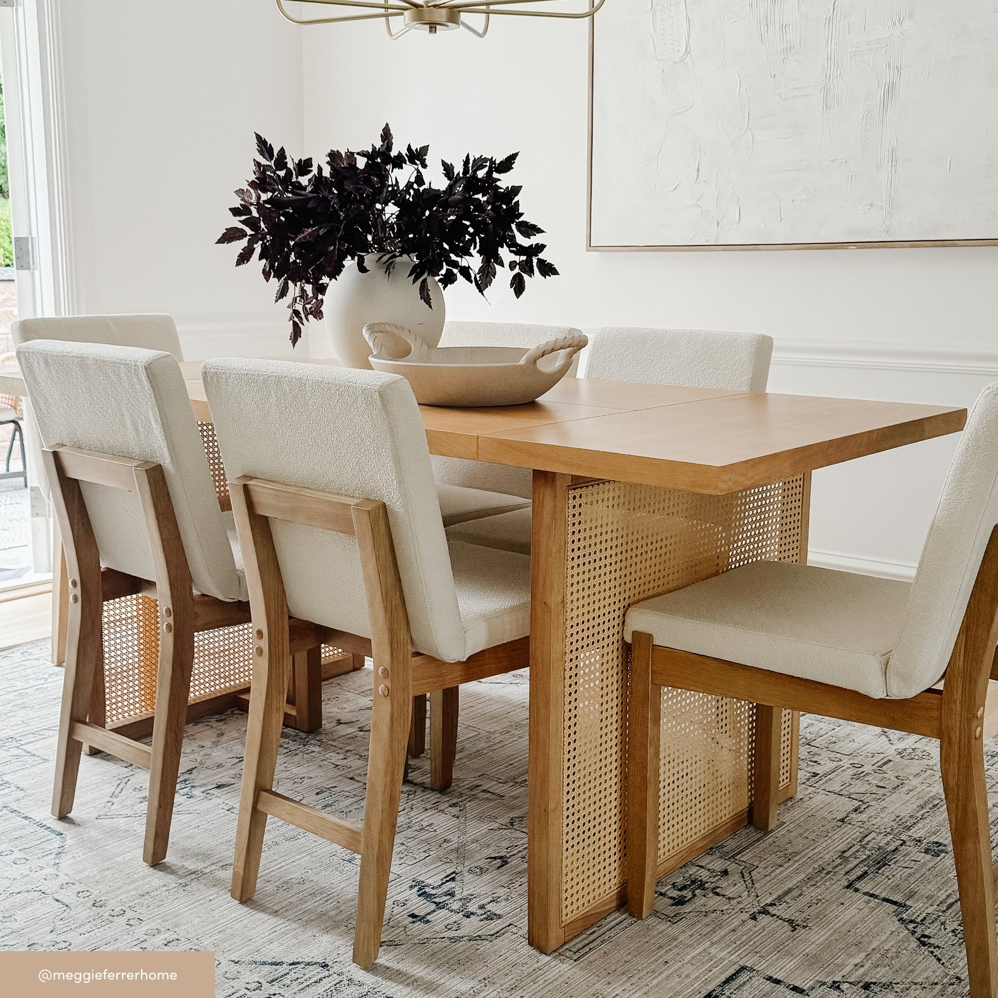 A modern dining room with a wooden table, six cushioned chairs with light fabric, a large ceramic vase with dark foliage, and a decorative bowl, all set on a patterned rug in a bright, white-walled space.