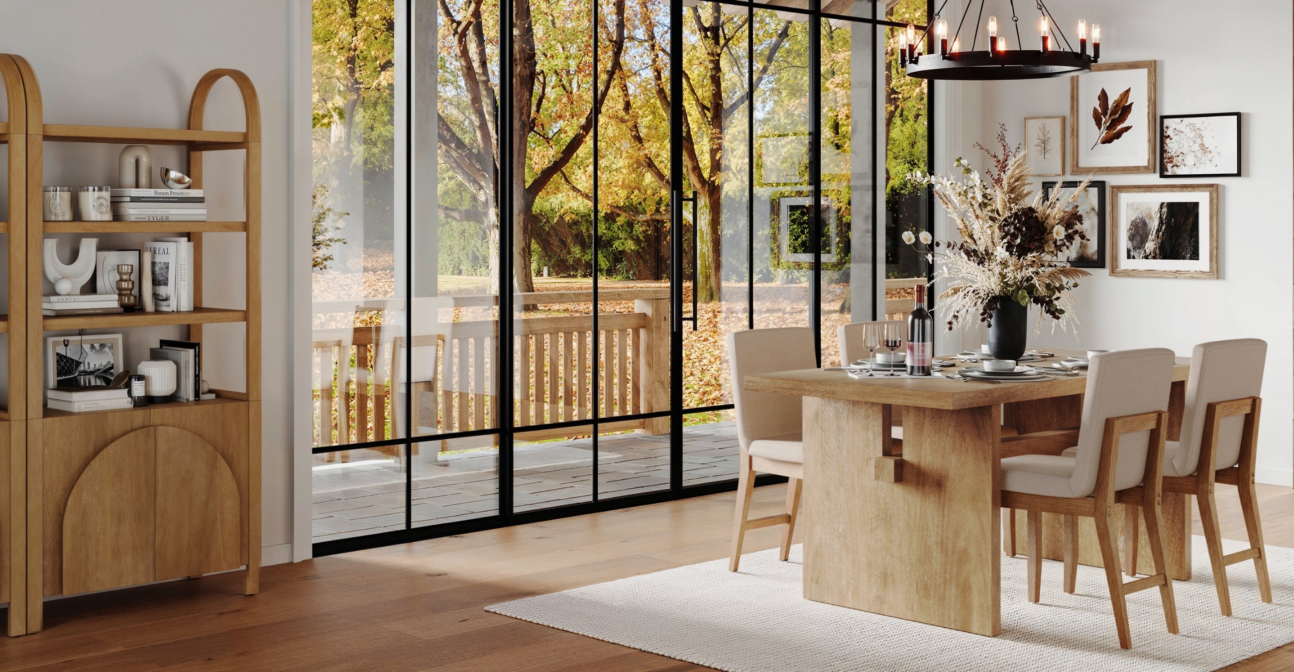 A modern dining room with light wood furniture, beige chairs, a set table, and a vase with dried flowers. Large glass doors reveal a deck and autumn trees outside. Shelves with decor and framed art hang on the white walls.