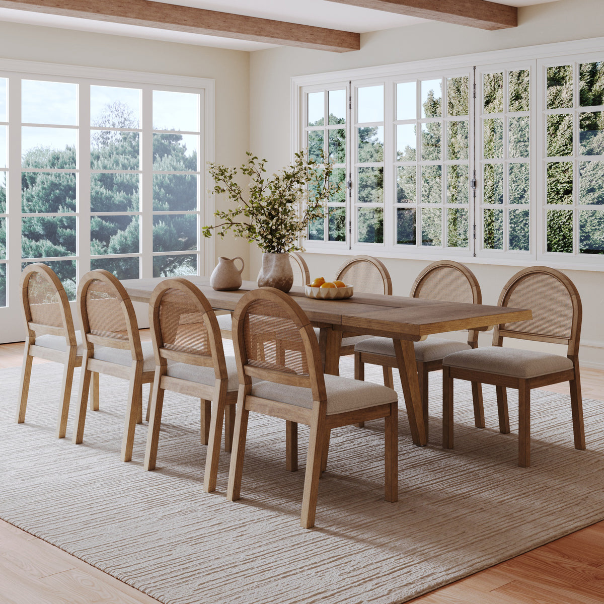 A wooden dining table with six matching chairs featuring cane backrests, arranged on a light-colored area rug in a sunlit dining room.