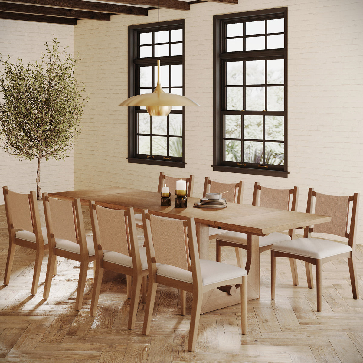 A dining room with a long wooden table, eight chairs with cane backs and light cushions, a potted tree, and a gold pendant light against a white brick wall.