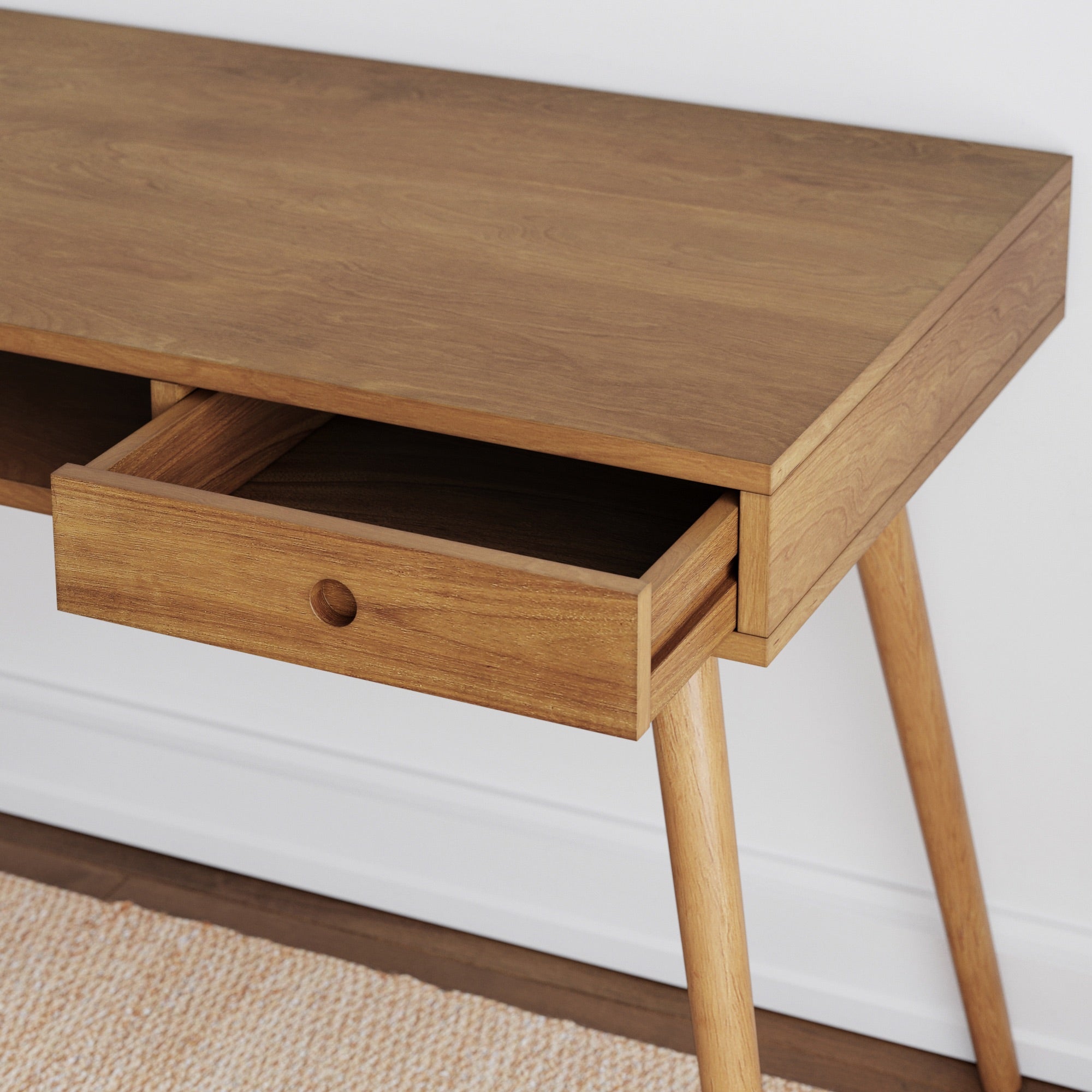 A wooden desk with angled legs and a single open drawer, placed against a white wall and above a beige textured rug.