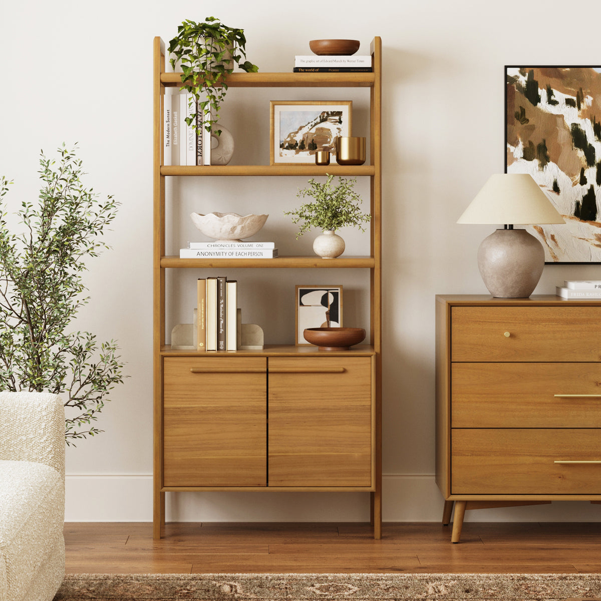 A wooden bookcase filled with books, decorative bowls, potted plants, and framed art, standing next to a matching three-drawer dresser topped with a table lamp.