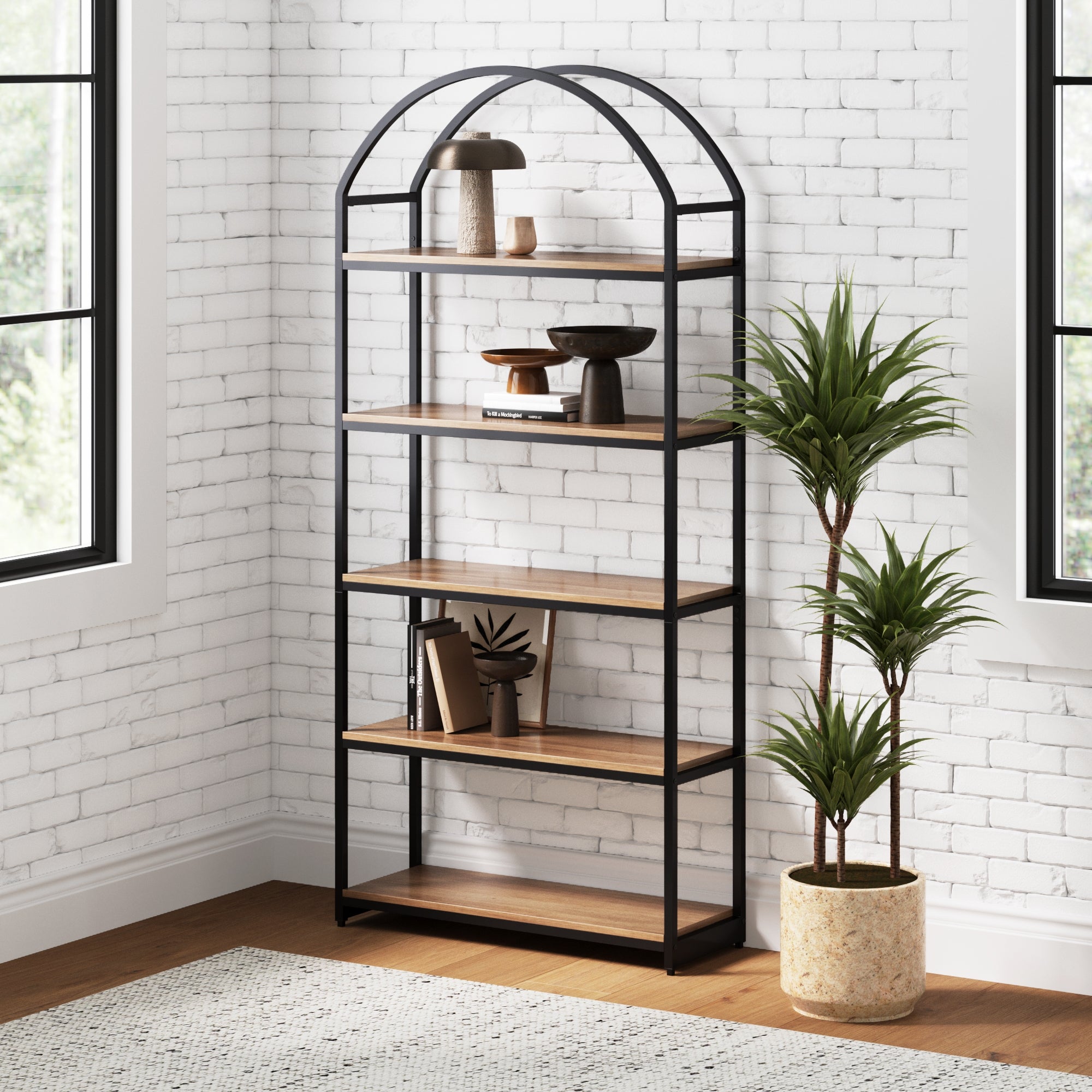 A black metal and wood bookshelf with an arched top stands against a white brick wall, styled with decorative objects. A tall potted plant sits beside it on a wooden floor near large windows.