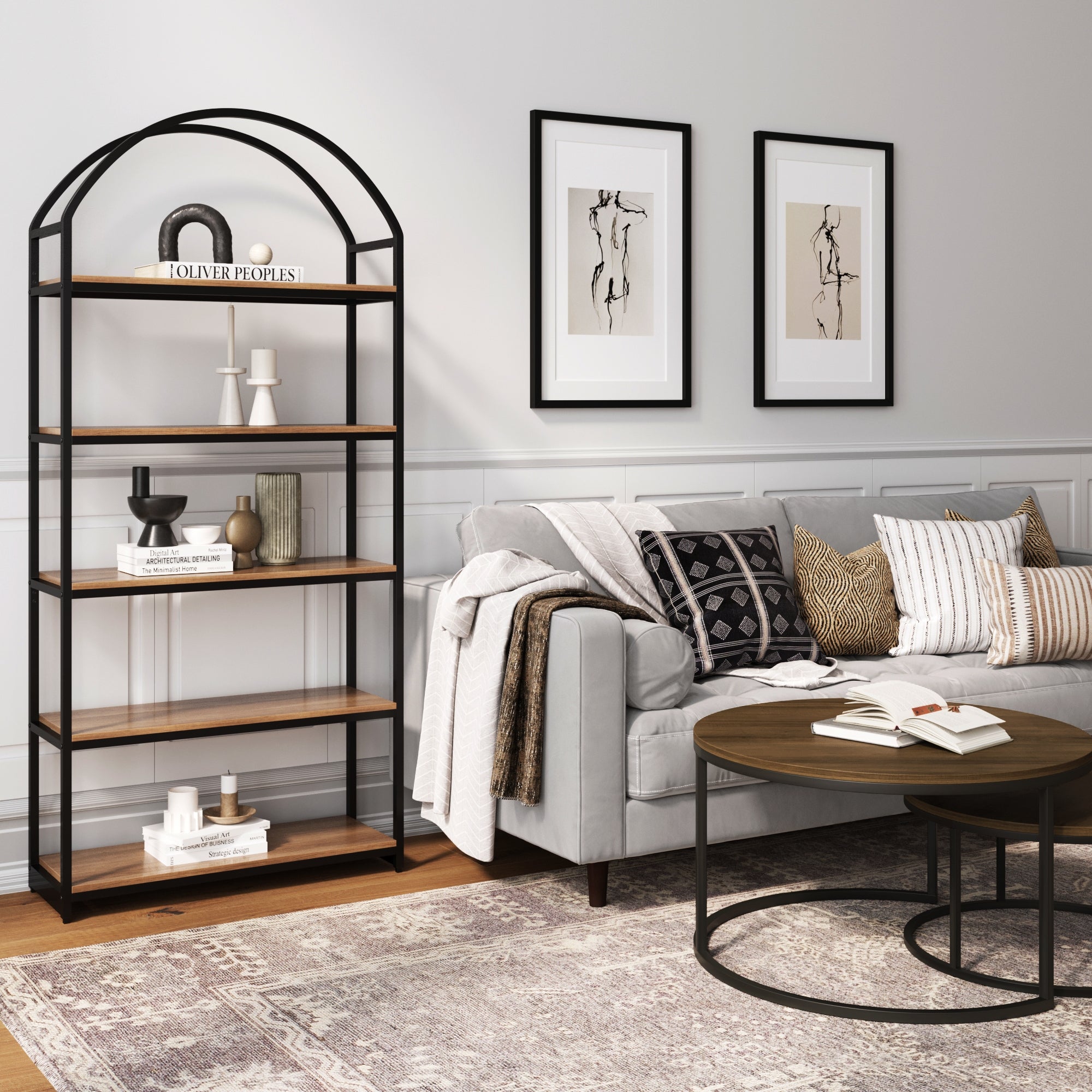 A modern living room with a light gray sofa, patterned pillows, round coffee table, and a tall, black-framed bookshelf with wooden shelves. Two abstract art pieces hang on the wall above the sofa.