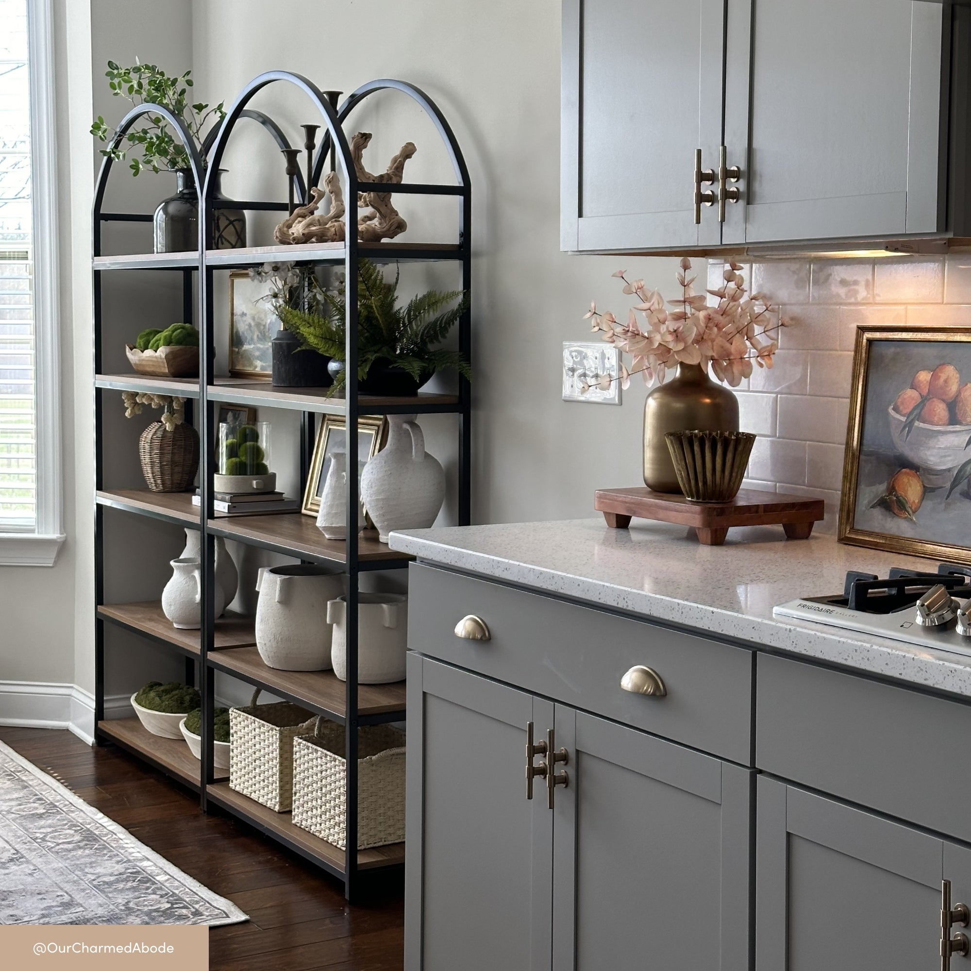 A modern kitchen with gray cabinets, a white countertop, and gold hardware. Open shelves hold baskets, plants, decor, and dishes. A vase with pink flowers sits on the counter near a framed painting of fruit.